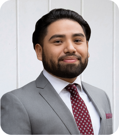 Professional man with dark hair and beard in a gray suit, white shirt, and patterned red tie, standing in front of a light-colored wall.