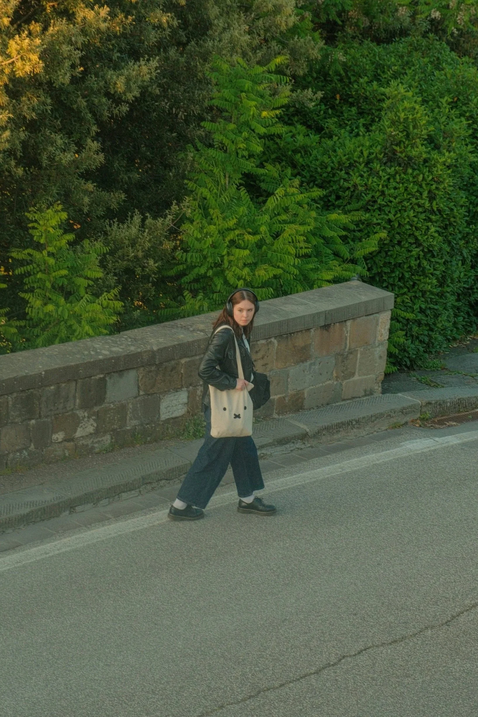 A woman crossing a street with a stone wall and green trees in the background.