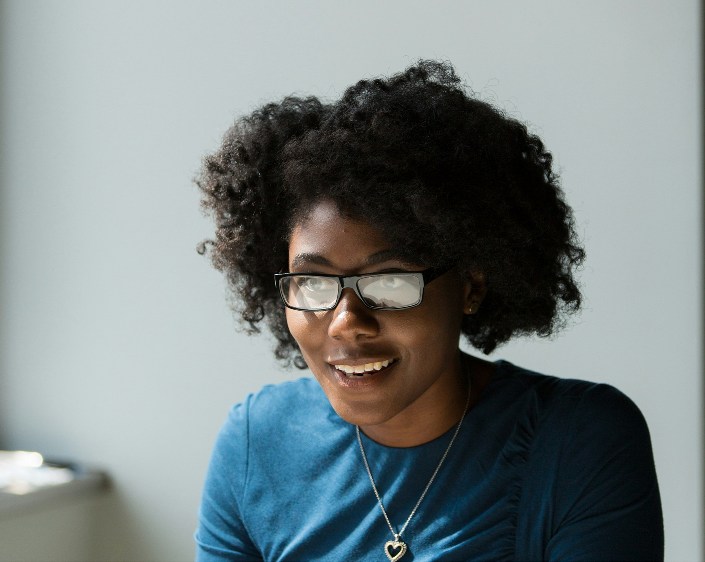 A woman with dark, curly hair wearing glasses and a blue shirt, smiling while looking down, with a blurred light background.