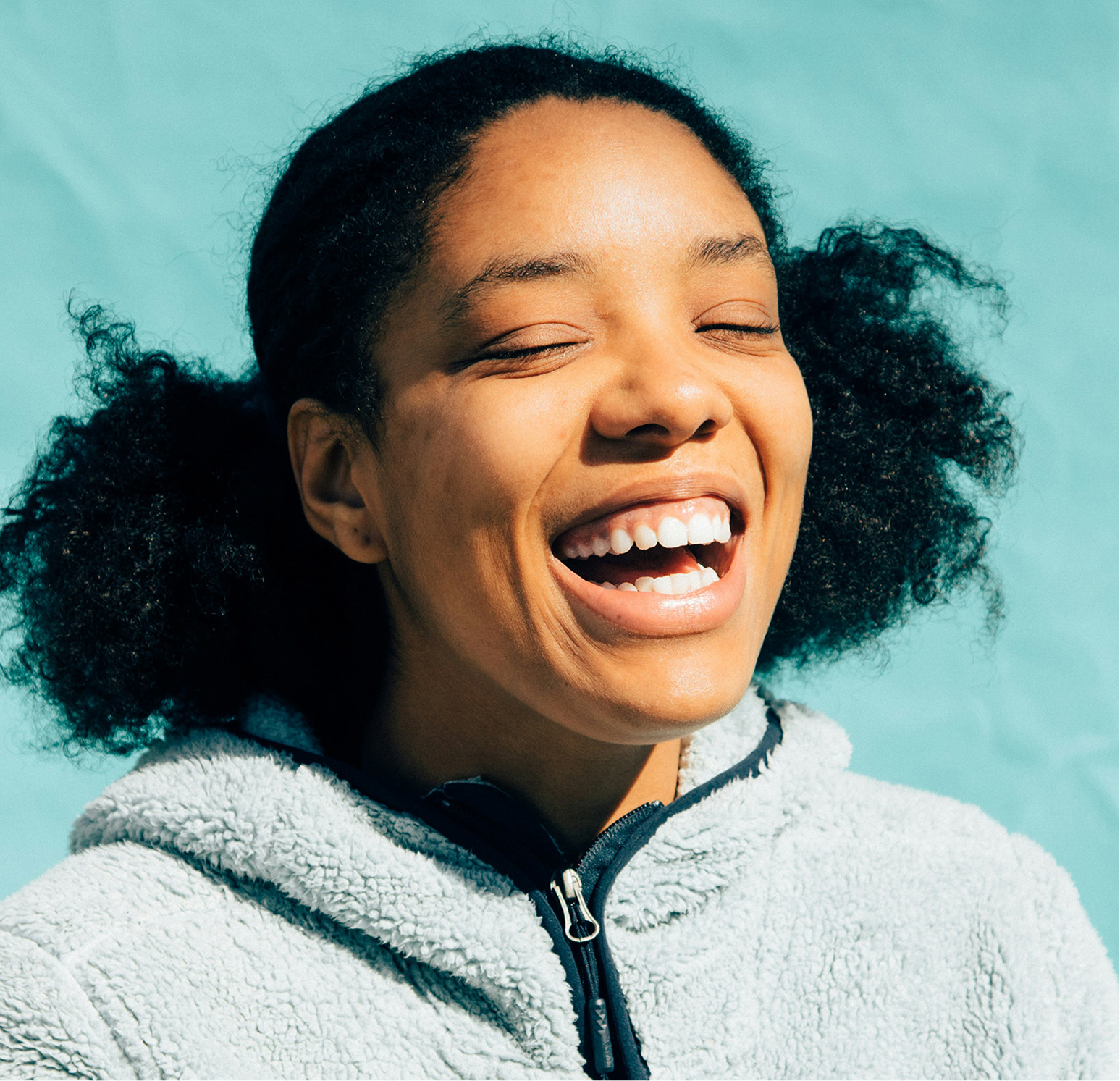 Close-up of a woman with curly dark hair laughing, wearing a white fleece jacket, against a light blue background.