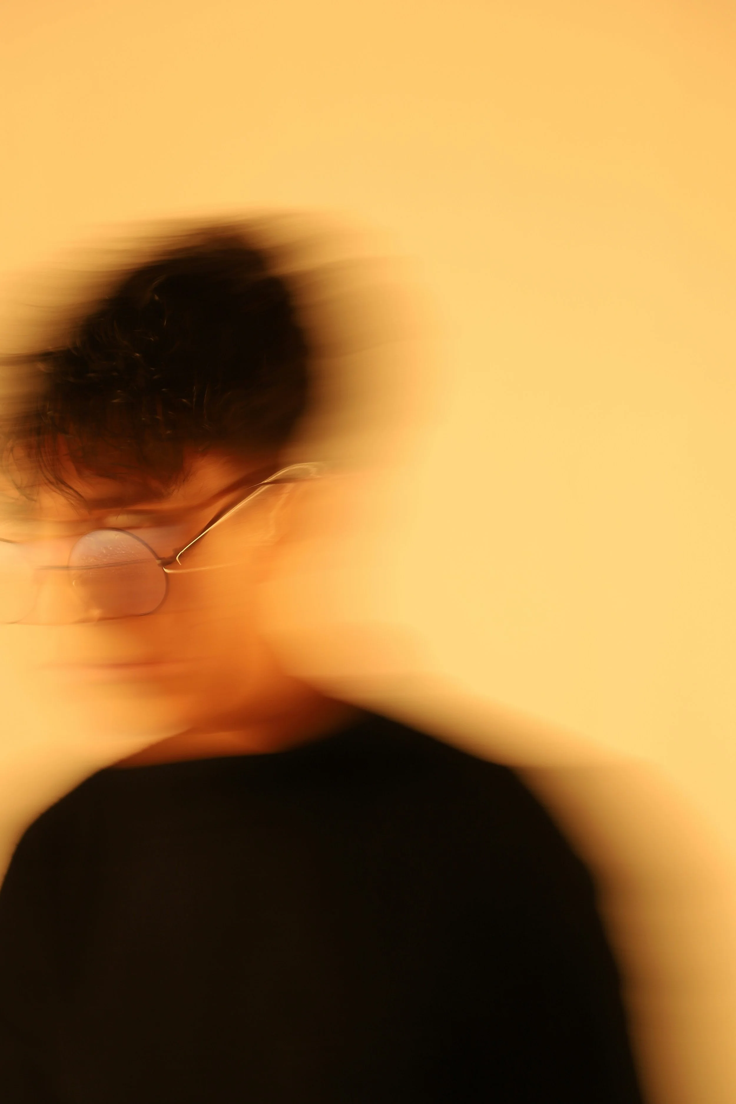 Person with glasses, blurred motion, dark hair, wearing a black shirt, against a yellow background.