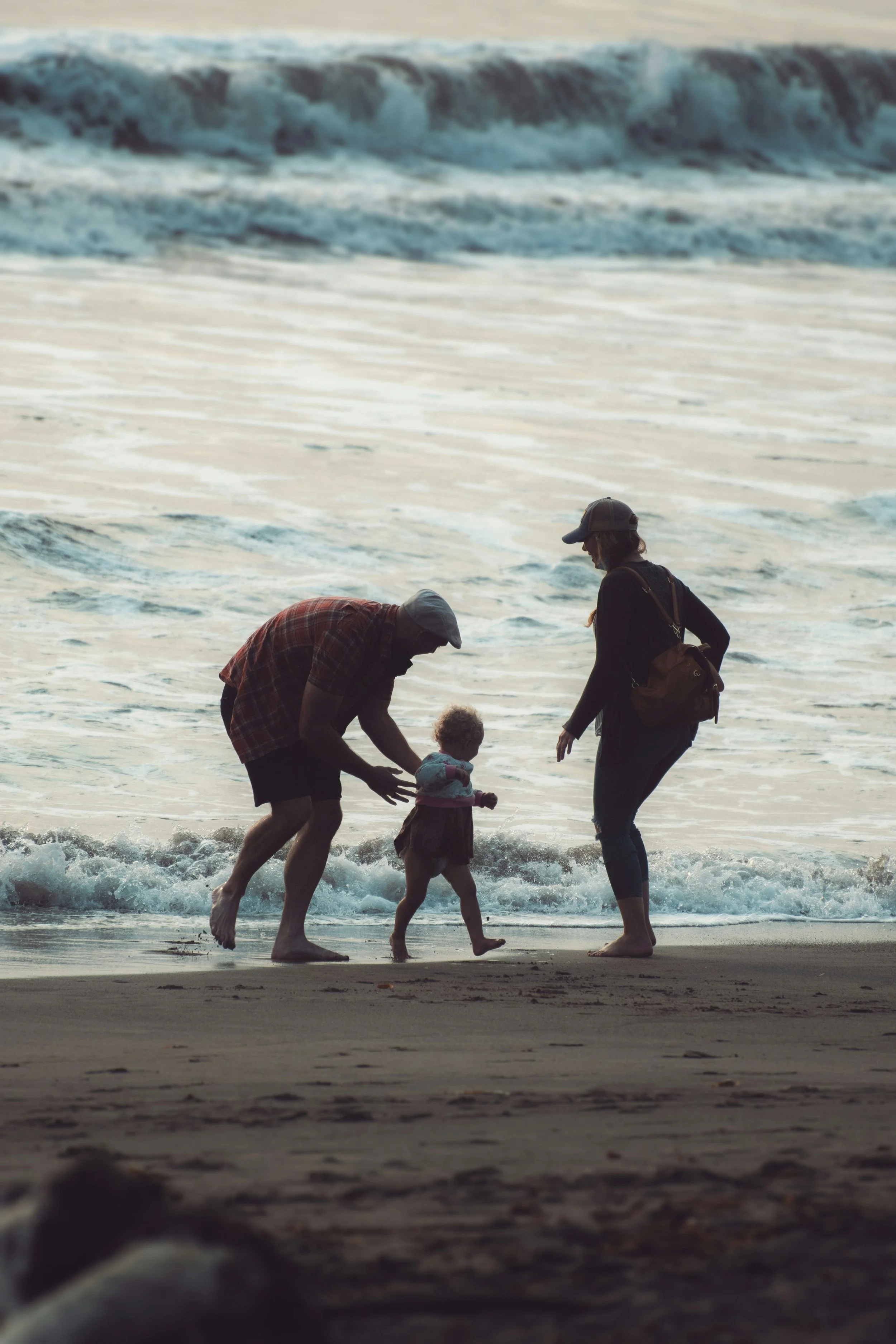 A man, woman, and young child at the beach, near the water's edge during sunset, with waves and ocean in the background.