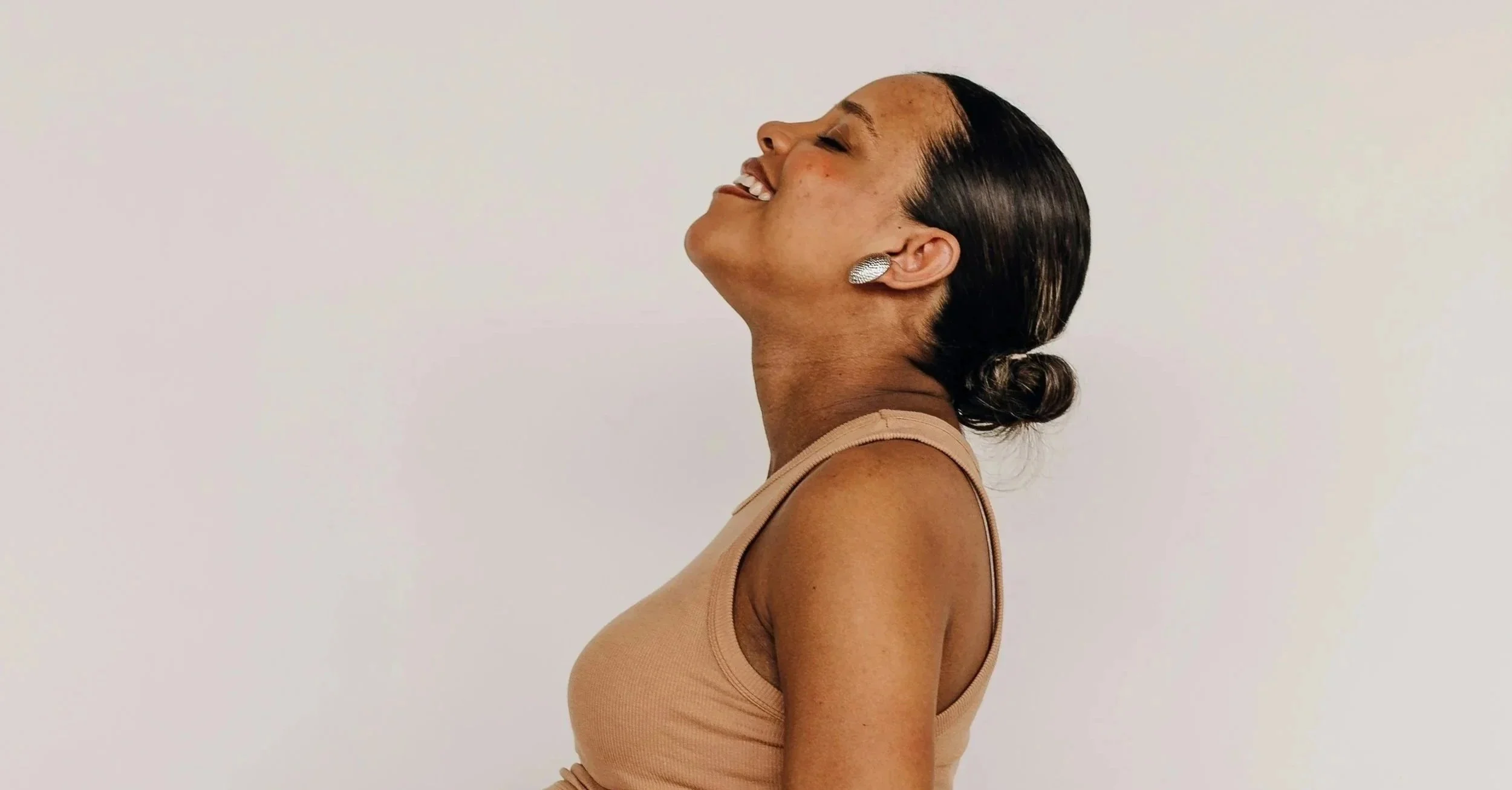 A woman with short, dark, wet-looking hair styled in a low bun, smiling with her eyes closed, wearing large, silver earrings and a beige sleeveless top, standing against a plain white background.