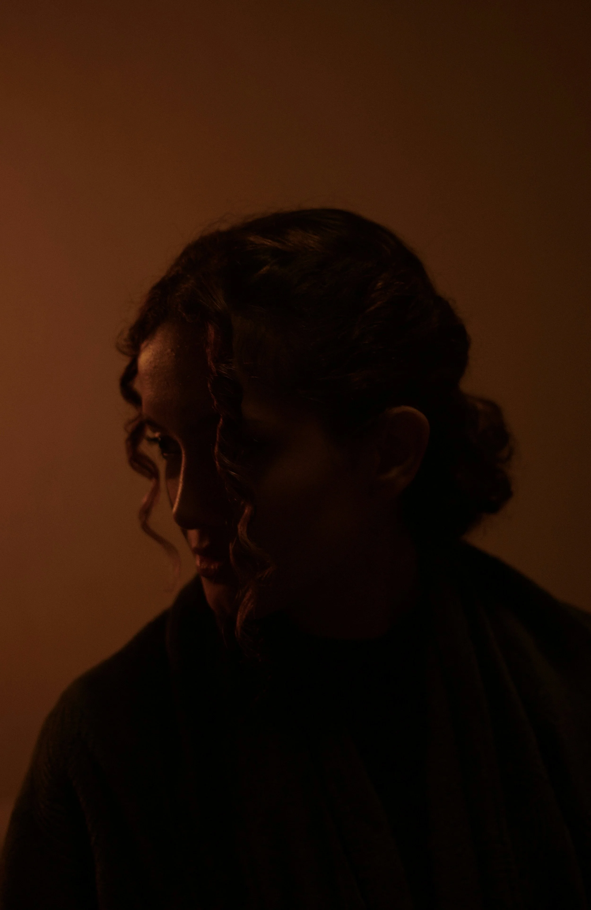A woman with dark hair styled in an updo and loose curls on the side, looking to the left in a dimly lit setting.