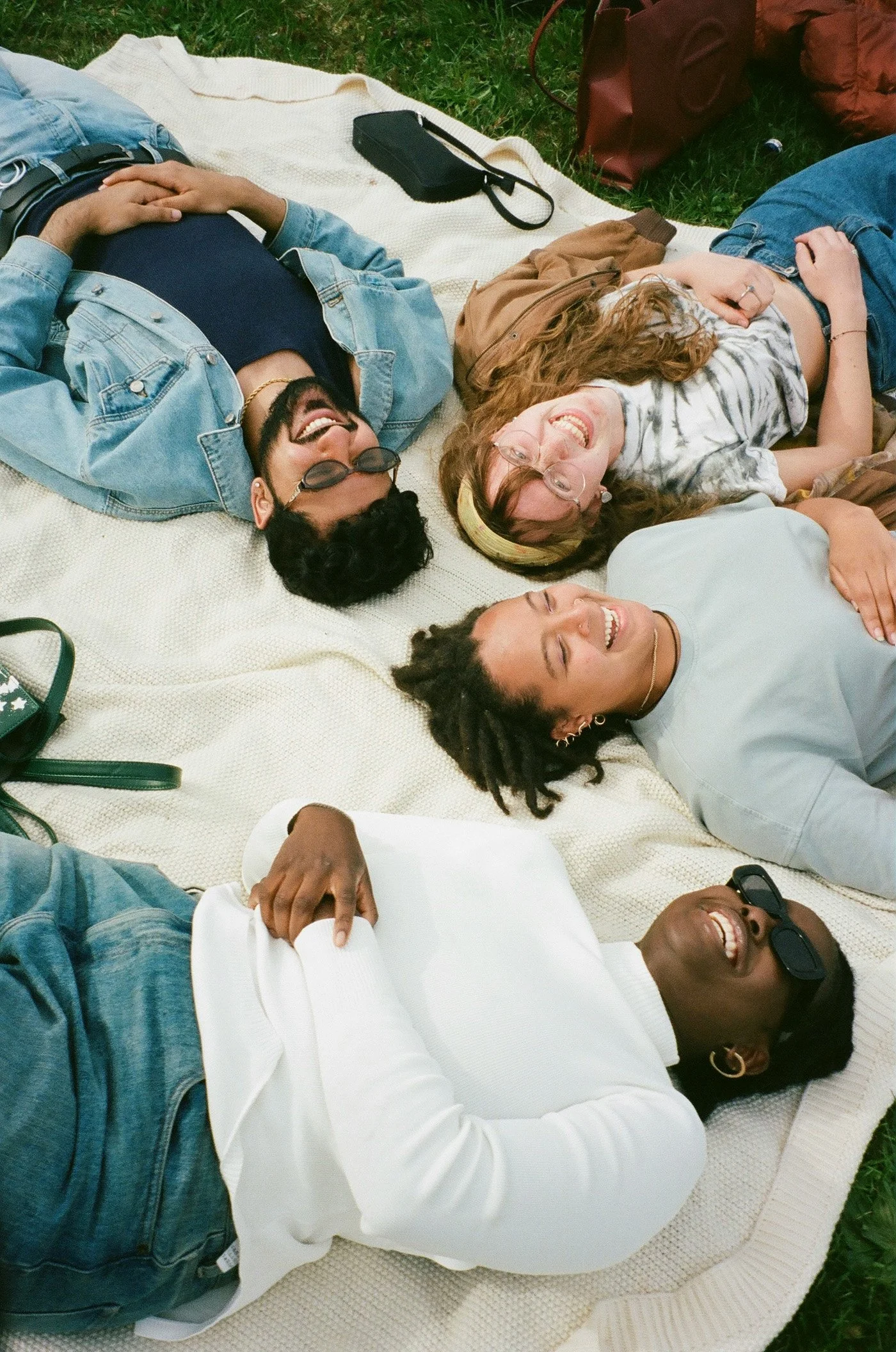 Five friends lying on a blanket in the grass, smiling and enjoying themselves outdoors.