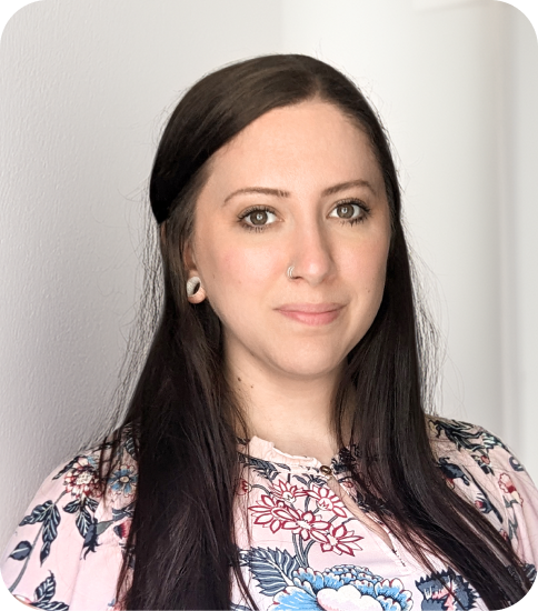 A woman with long dark hair, wearing a floral-patterned blouse and nose ring, standing indoors against a plain wall.