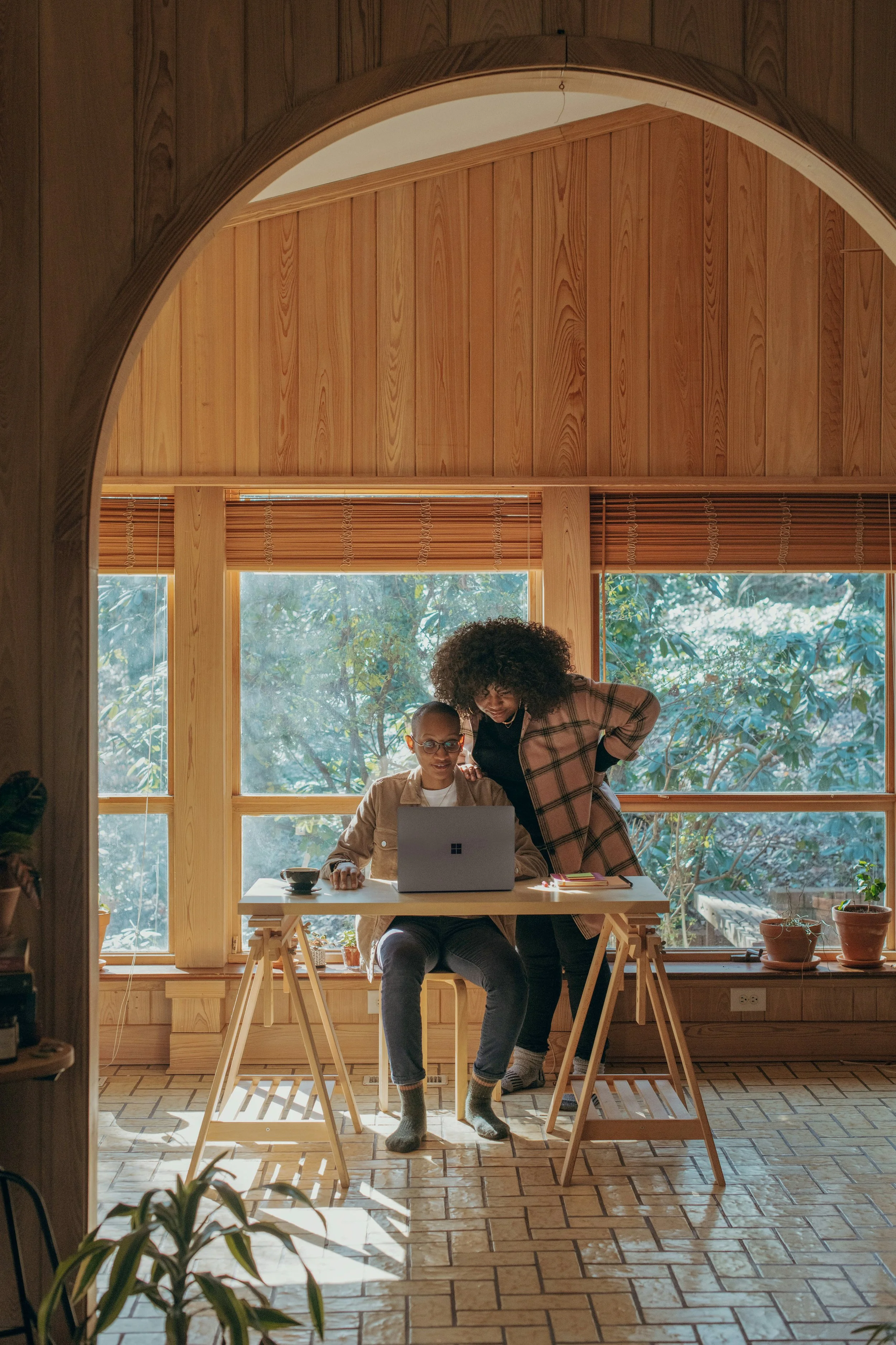 Two women are looking at a laptop in a bright room with large windows and wooden interior, sitting at a wooden desk.