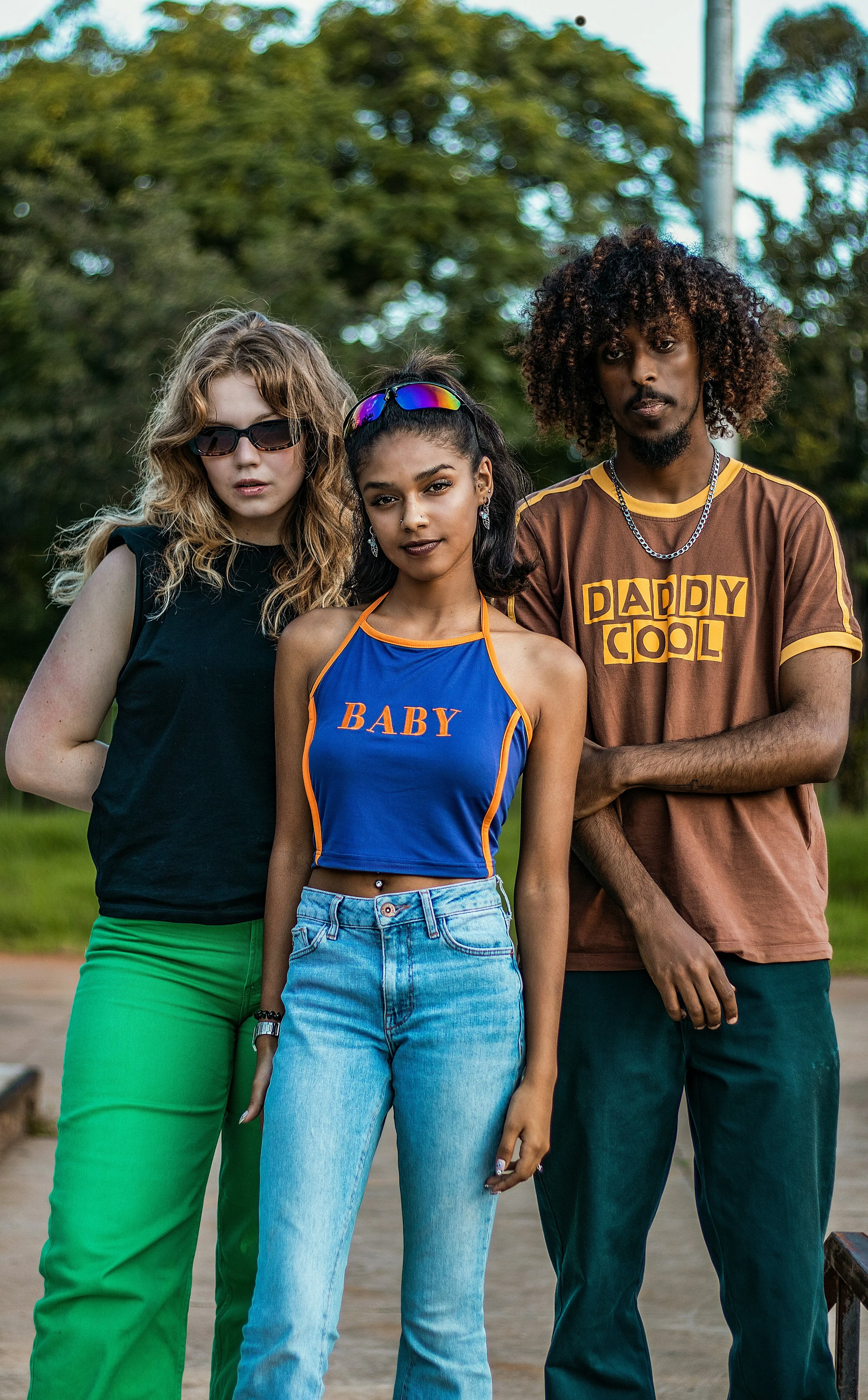 Three young people standing outdoors, a woman with curly blonde hair and sunglasses, a woman with long black hair and reflective sunglasses, and a man with curly hair and a brown t-shirt that says 'DADDY COOL',
