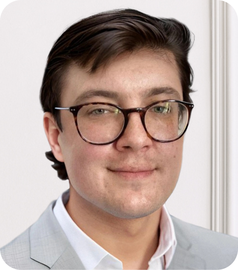 A young man with brown hair, glasses, and a slight smile, wearing a light gray blazer over a white shirt, posing indoors.