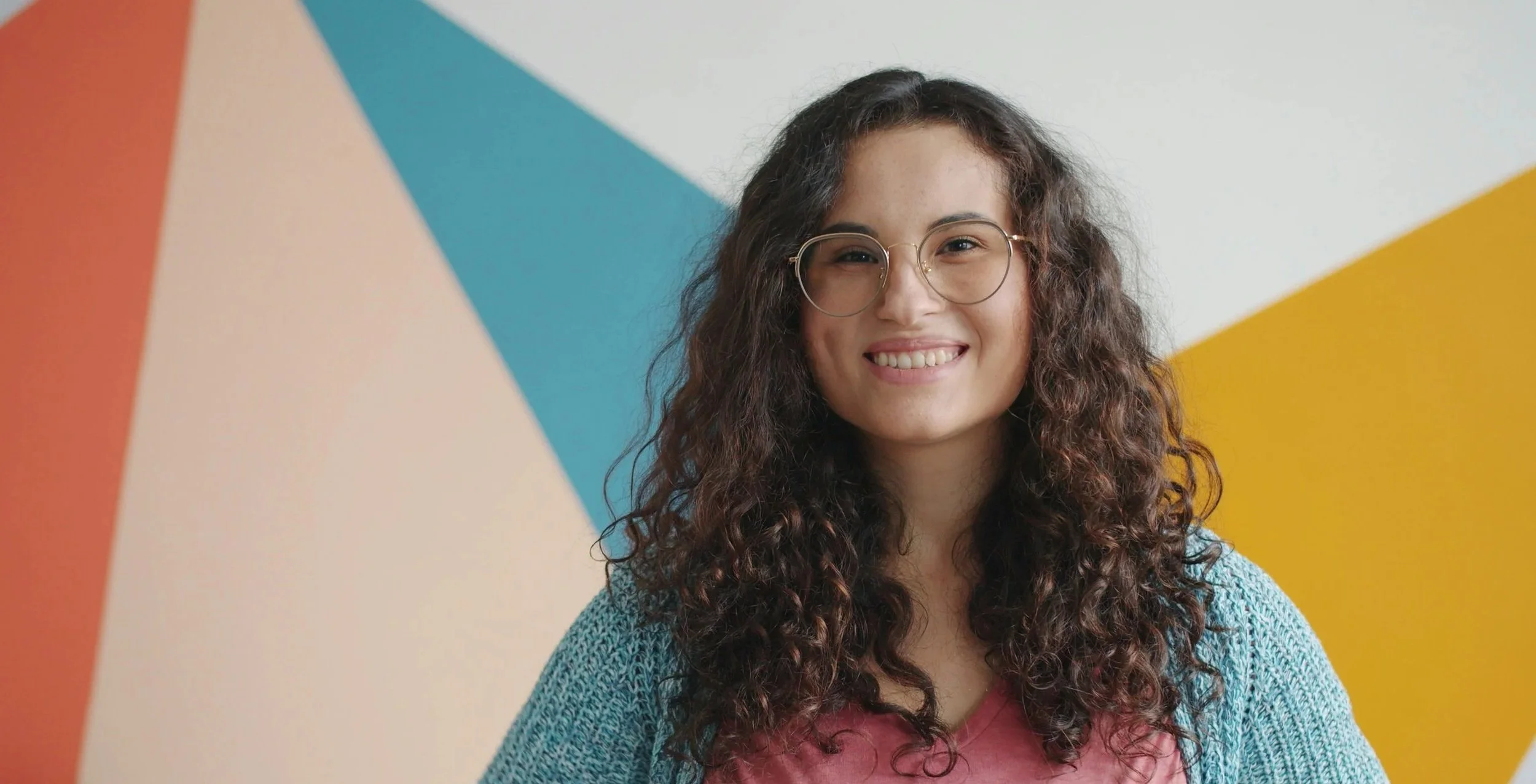 A young woman with long curly brown hair and glasses smiling, wearing a pink shirt and a blue sweater, standing in front of a colorful geometric wall with sections of orange, beige, blue, and yellow.