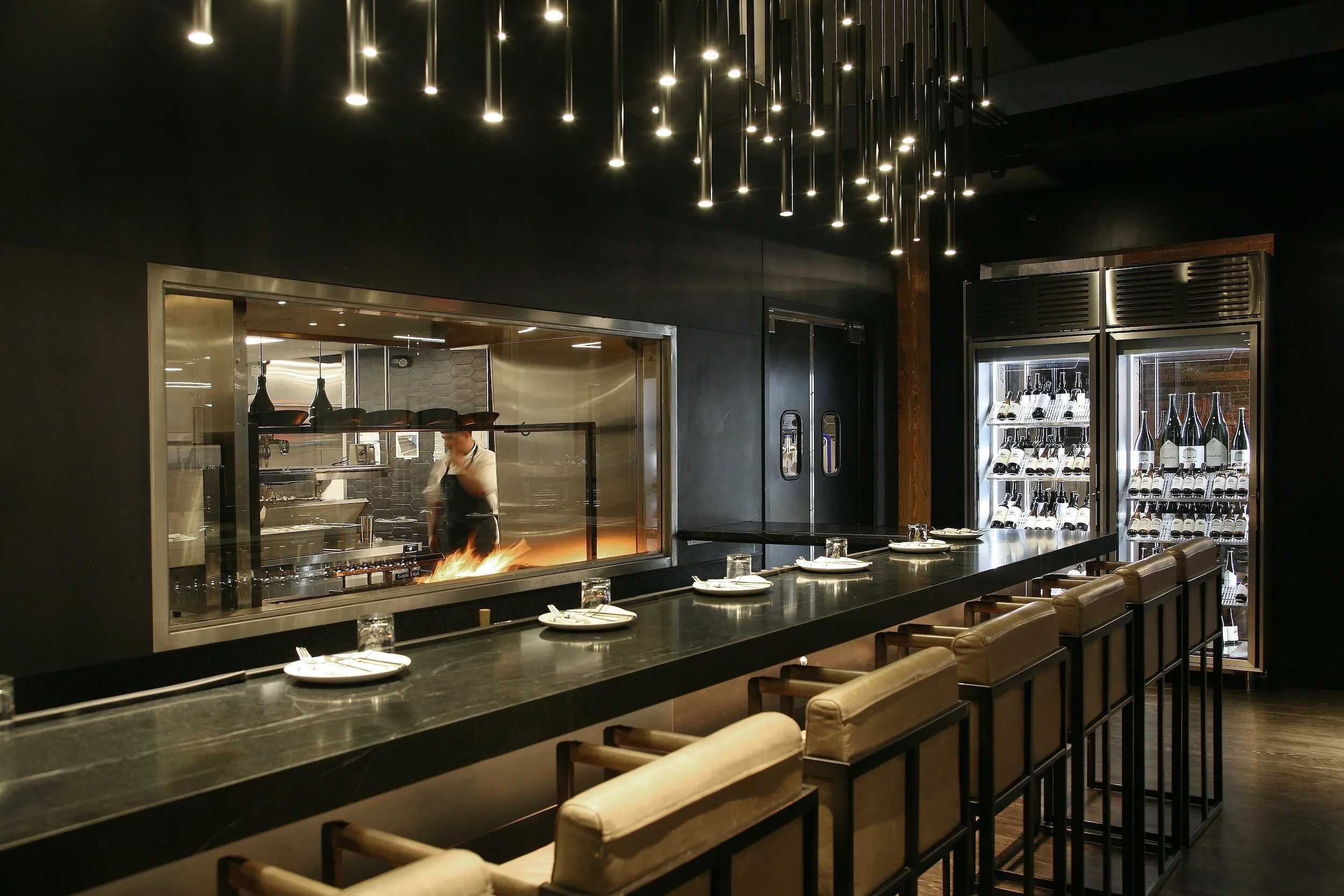Modern restaurant interior with a bar counter, high-backed chairs, and a view into the open kitchen where a chef is cooking. Wine bottles in a refrigerator are visible in the background, and the ceiling features hanging lights.