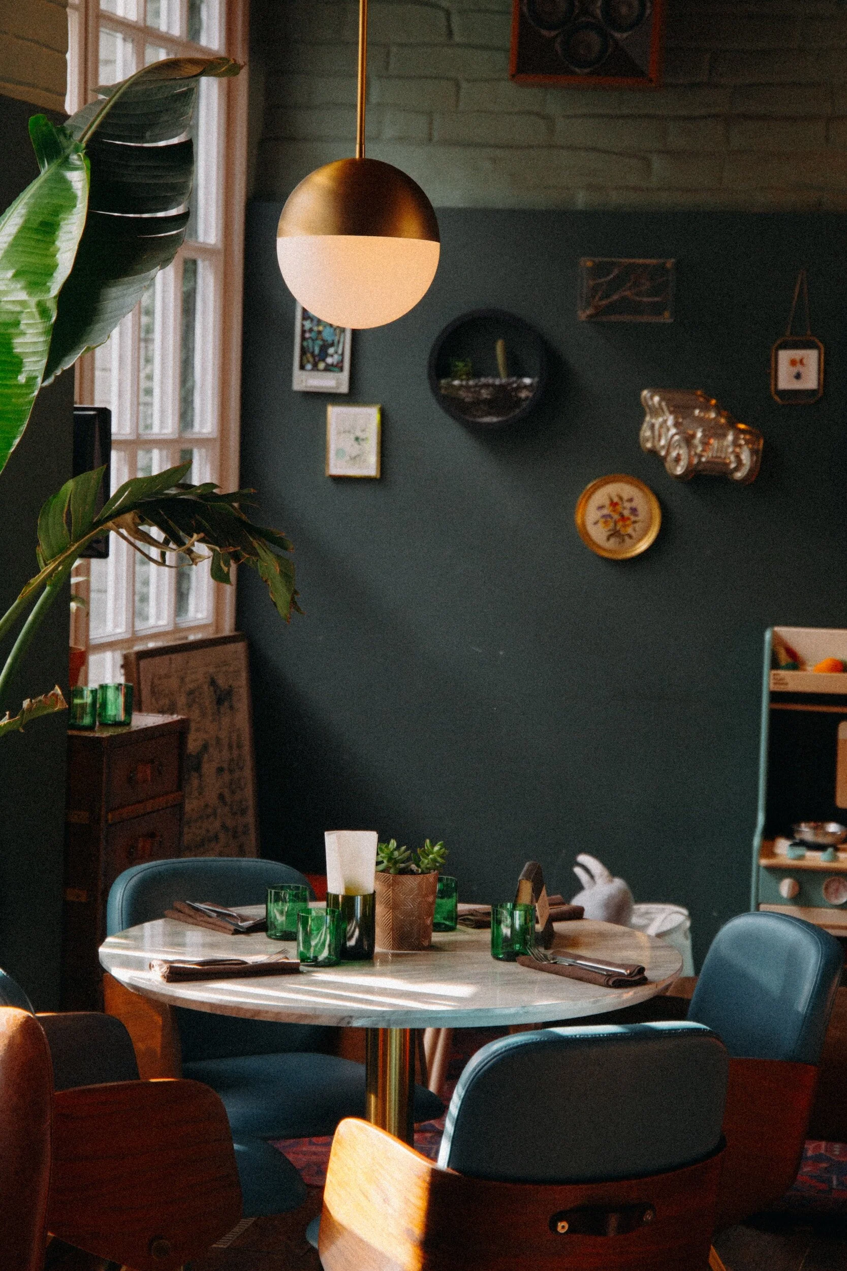 A cozy dining area with a round marble table, surrounded by teal and wooden chairs, decorated with green glasses and a small plant in the center. A large window lets in natural light, and a modern hanging light fixture is above the table. The dark green wall is decorated with various framed art pieces and decorative objects, and a large leafy plant is visible on the left side of the image.