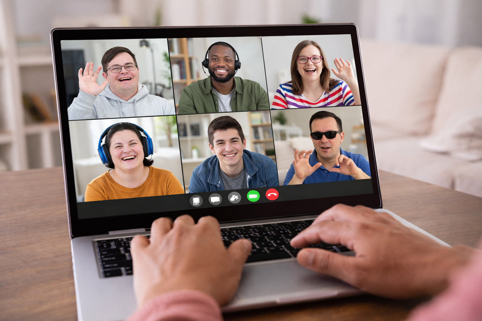 Person using a laptop to participate in a video conference call with six diverse smiling people, some waving, in a well-lit room.