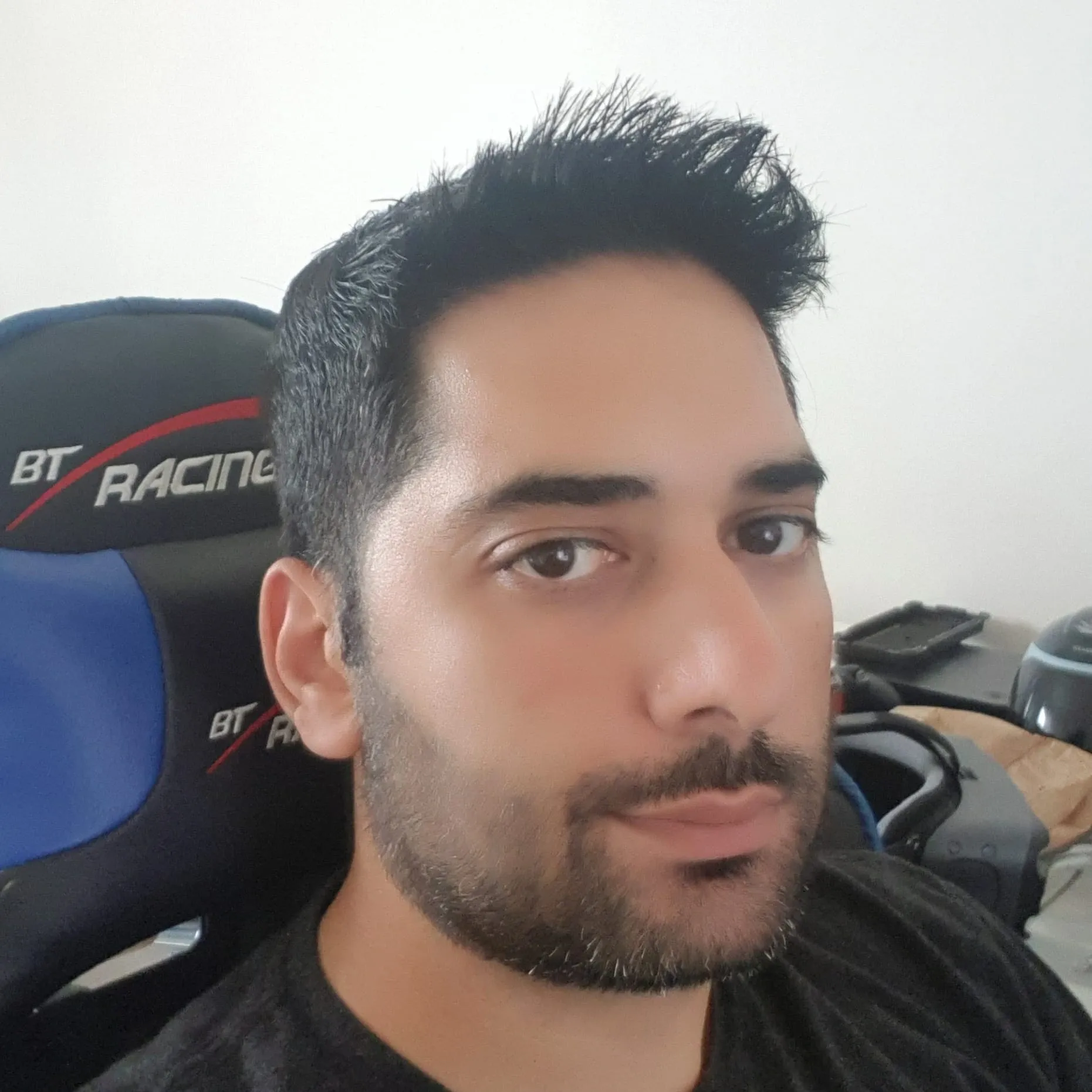 Close-up of a man with dark hair and facial hair sitting in a room with a white wall, a black and blue gaming chair with 'BT Racing' logo, and various items on a desk in the background.