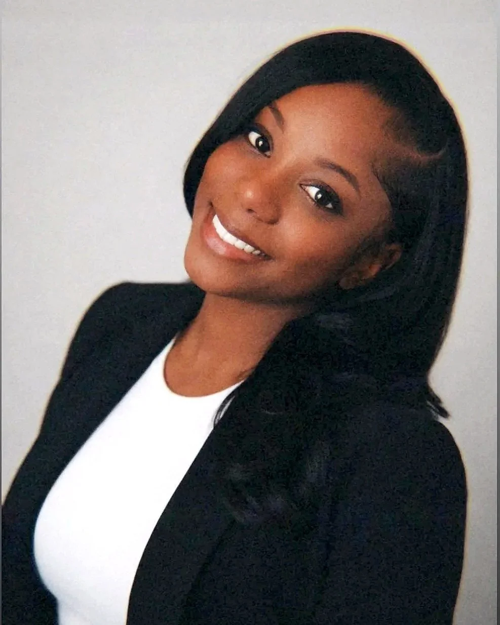 Portrait of a young African American woman with long black hair, smiling, wearing a black blazer over a white top, against a neutral background.