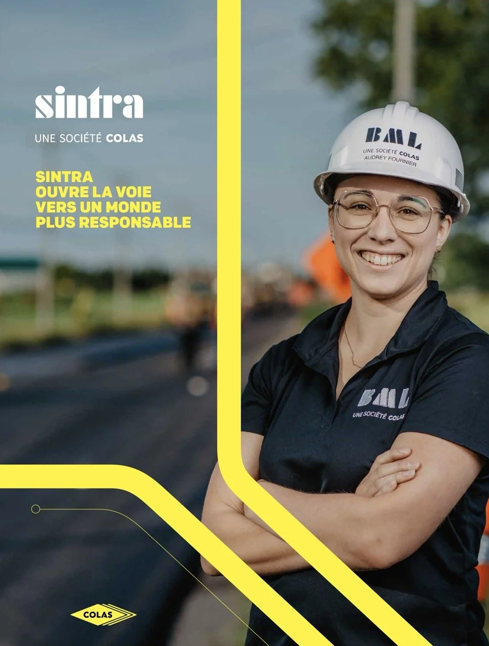Une femme portant un casque de sécurité blanc avec le logo de COLAS et un uniforme noir, souriante, devant un chantier de construction routière en plein jour. Conception graphique par l'agence de communication Niveau 5, situé dans Bellechasse.