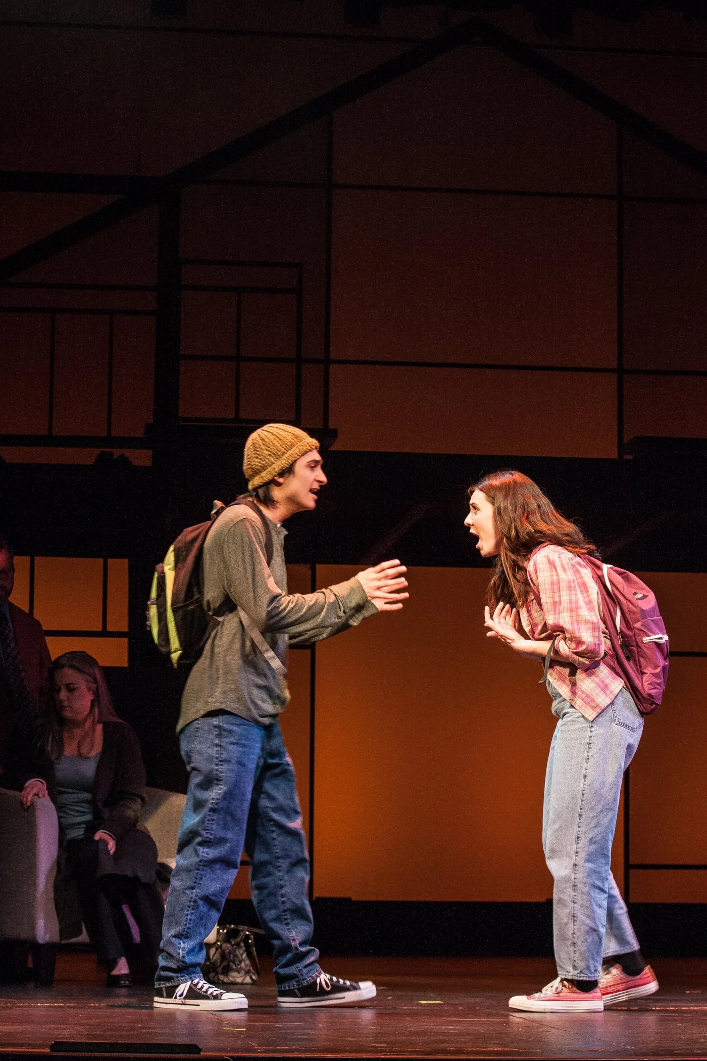 Two young actors perform on stage, facing each other intensely, with one gesturing with hands and the other leaning forward, wearing casual clothing and backpacks. In the background, three seated individuals observe the scene.