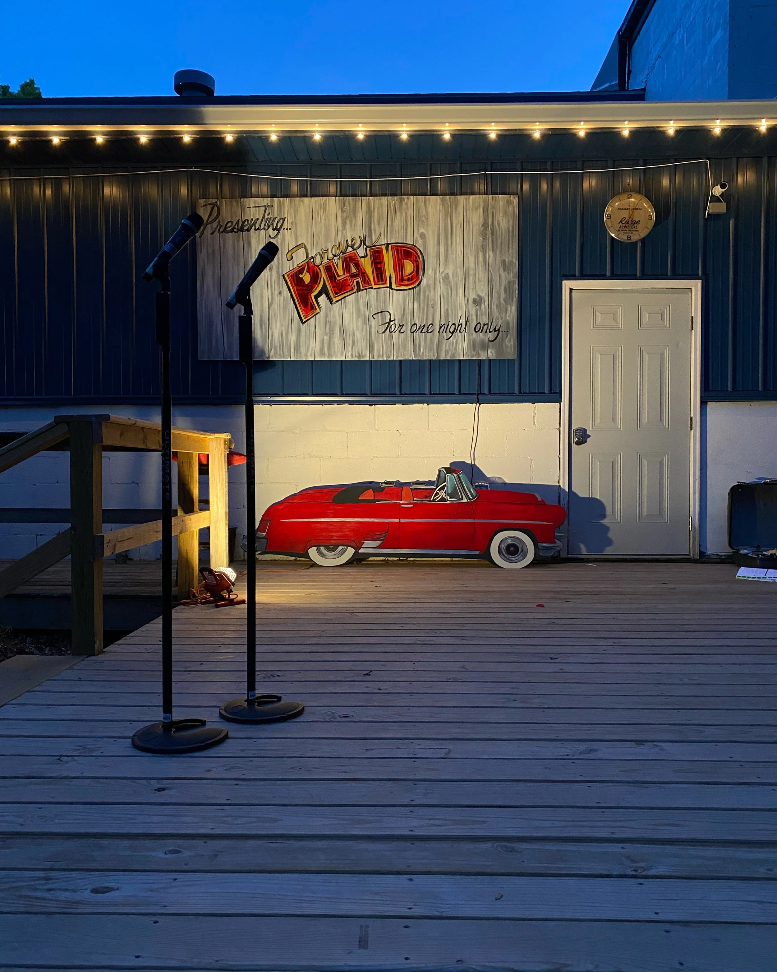 Stage setup on an outdoor wooden deck with two microphones, a red vintage car-shaped cutout, and a sign reading 'Power PLHAD For one night only' on the building wall. Blue sky in the background.