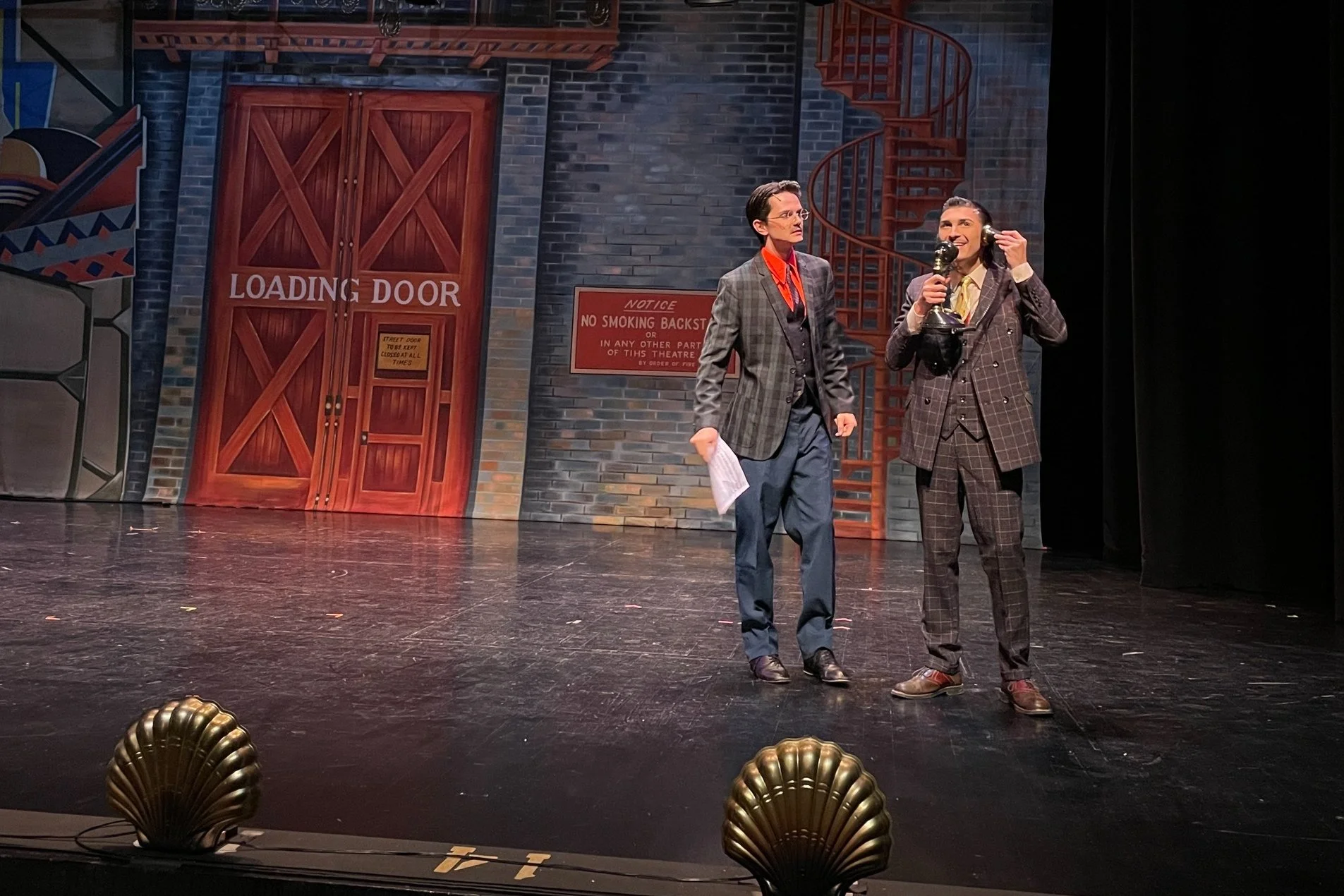 Two actors on stage, dressed in vintage suits, one holding a telephone, with a painted backdrop resembling a city alley and a loading door, stage lights in the foreground, during a theatre performance.