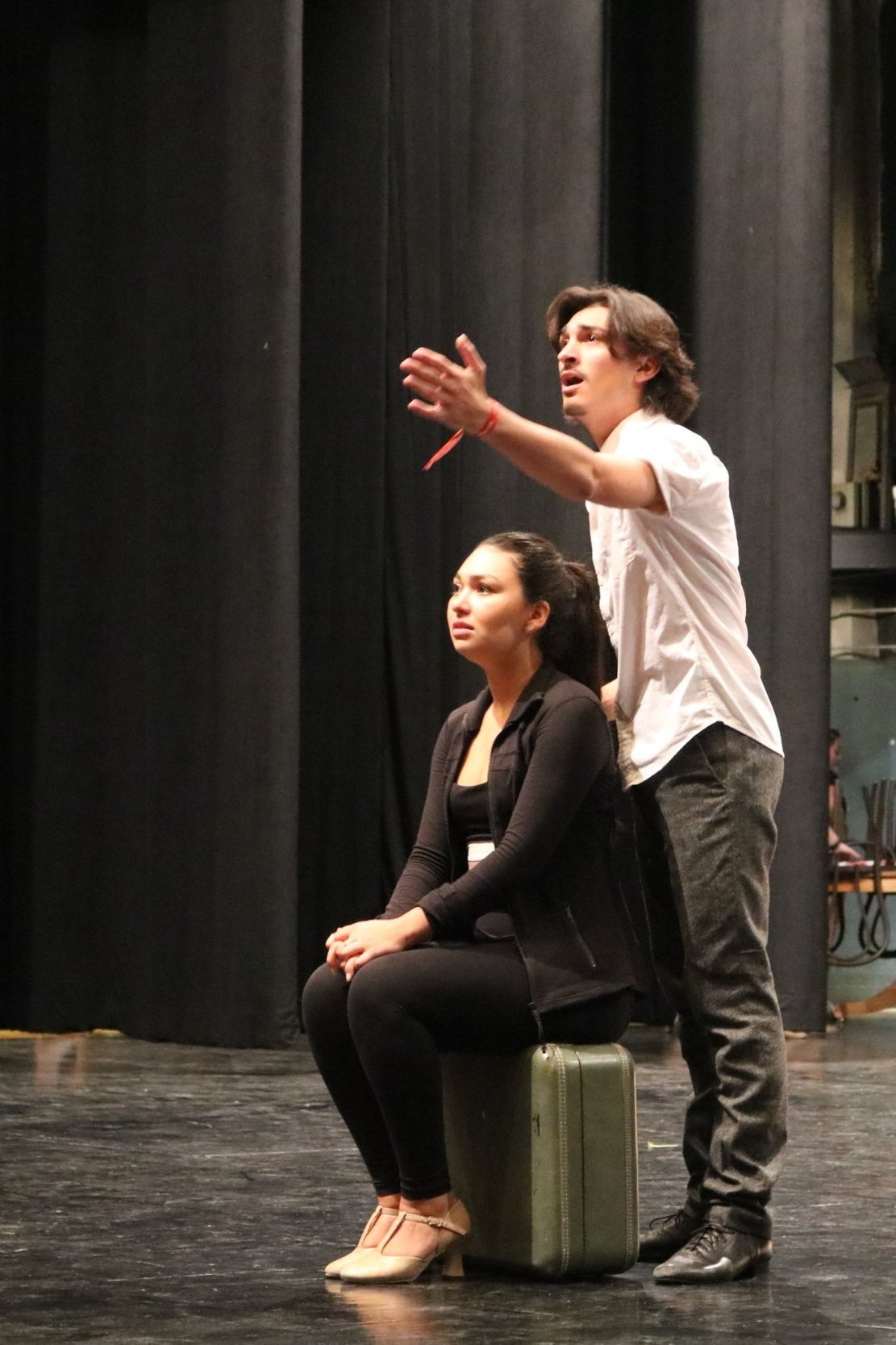 A man and a woman on stage during a theatrical performance. The woman is sitting on a suitcase, wearing black, and the man is standing beside her, gesturing with his hand. The background has black curtains.