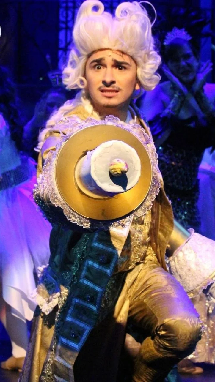 Person dressed as a regal character in an elaborate costume with a large white wig, holding a gold tray with a hat and cake, on stage with other performers in the background.