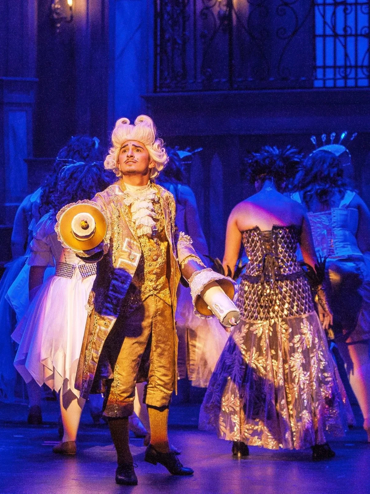 A performer dressed in elaborate historical costume, holding a hat, performing on stage with other performers in ornate dresses behind him.