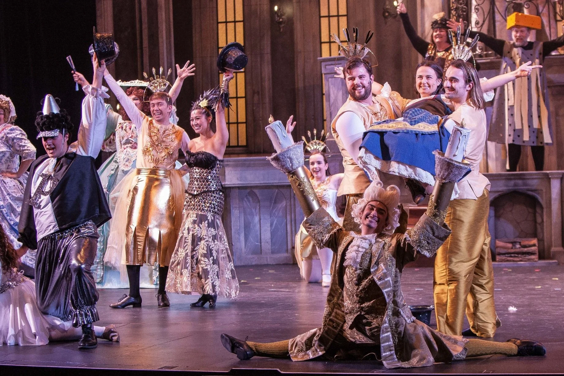 Actors in a theatrical production dressed in elaborate costumes, celebrating on stage with joyful expressions and raised arms.