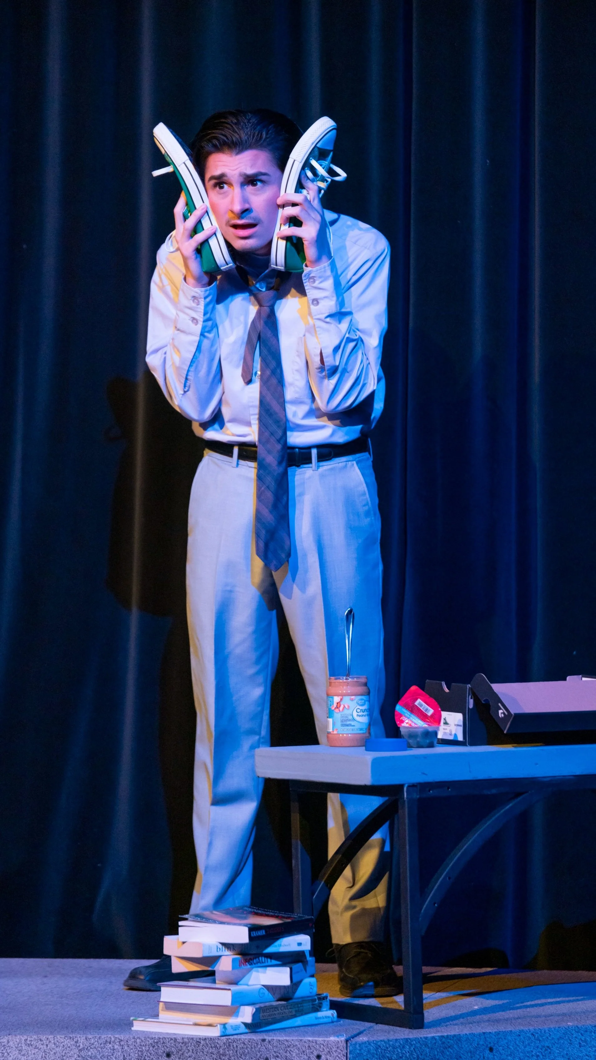 A man with dark hair and wearing a light blue shirt, gray pants, and a gray tie, holding a pair of green and white sneakers to his ears as if they were a phone, standing on a stage with a black curtain backdrop. There is a table with books, a jar of 