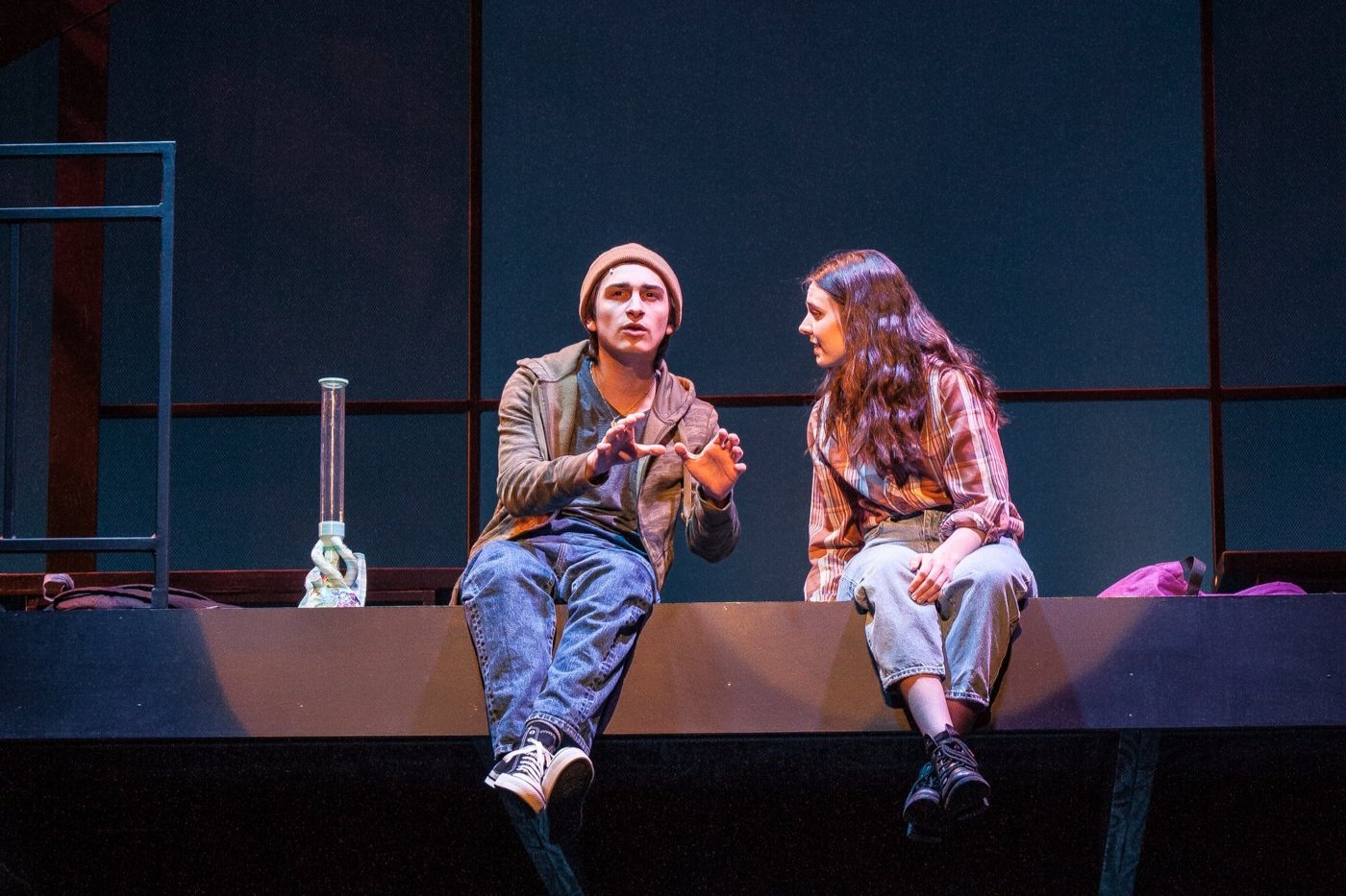 Two young actors sitting on the edge of a stage, engaged in conversation, with a science beaker in front of them.