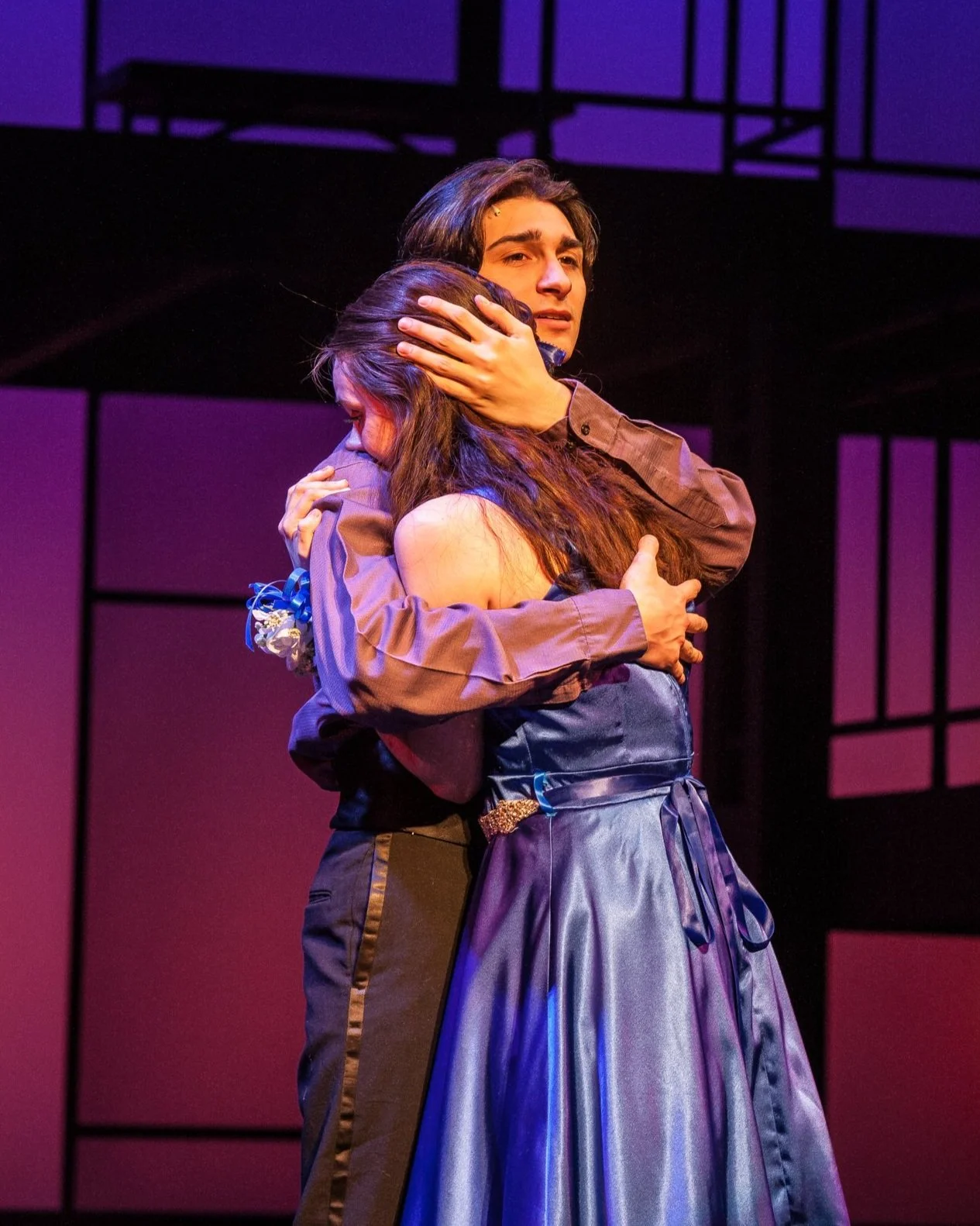 A young man and woman sharing an emotional hug on stage, with the man holding the woman's head and the woman with her face pressed against his chest, lit with purple and blue stage lighting.
