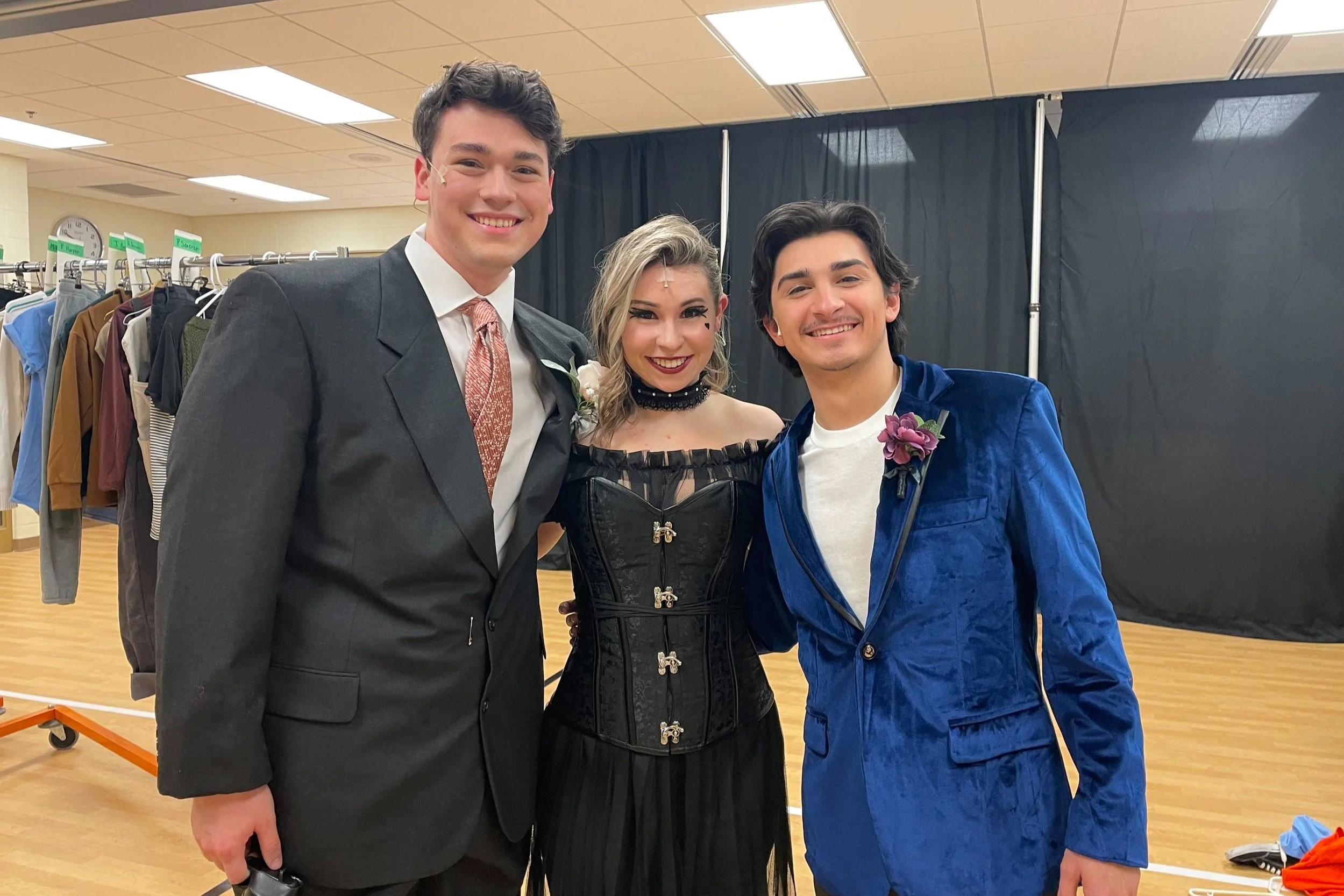 Three young adults dressed in fancy costumes, smiling and standing together in a room with racks of clothes.