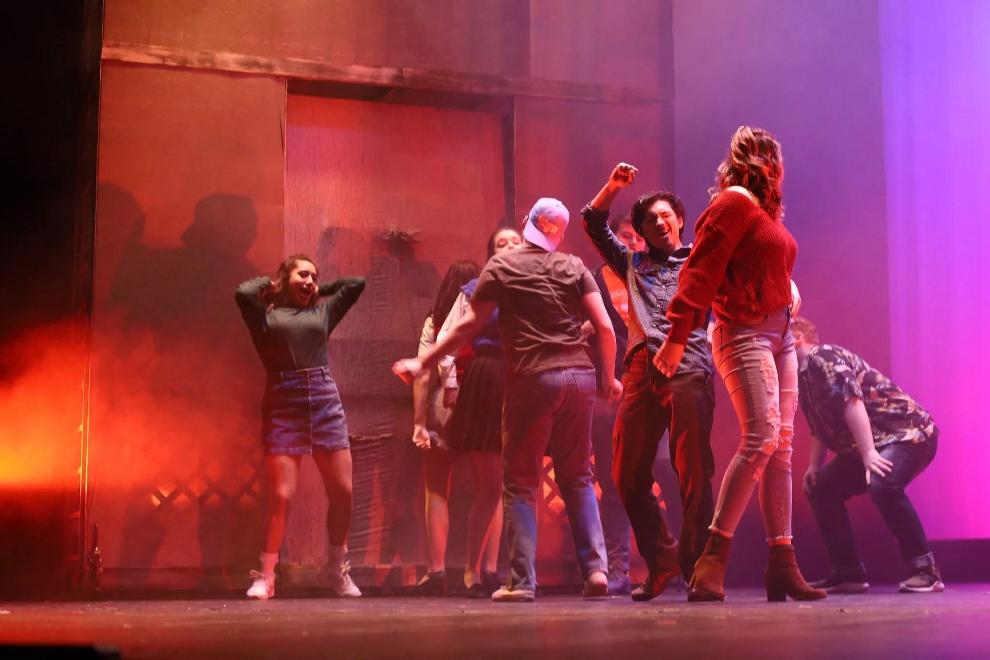 Group of young people dancing and celebrating on stage with colorful lights, some with raised fists, enjoying a lively performance.