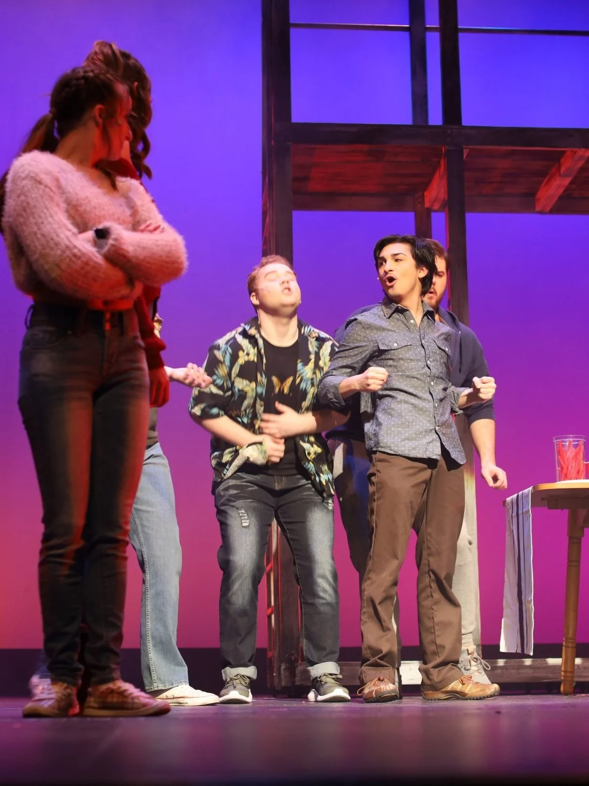 People performing on stage, with one person in the foreground with their arms crossed, and a group of others singing or speaking passionately.