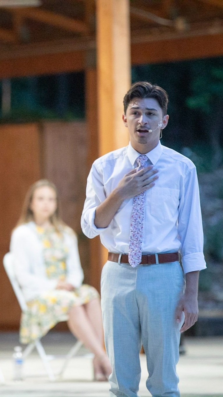 A man in a white shirt and light-colored pants with a floral tie, speaking and touching his chest, while a woman sits on a white chair in the background. The scene appears to be a performance or talk in an indoor or semi-outdoor setting.