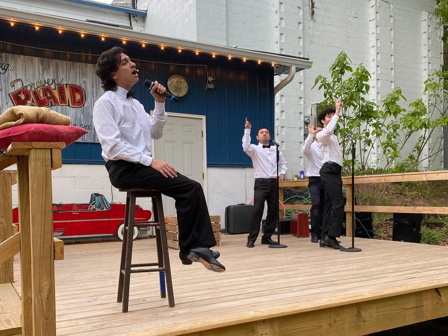 A group of four performers dressed in black pants and white shirts with black bow ties, performing on an outdoor wooden stage. One person is sitting on a stool, singing into a microphone, while three others stand behind, with two singing into microph