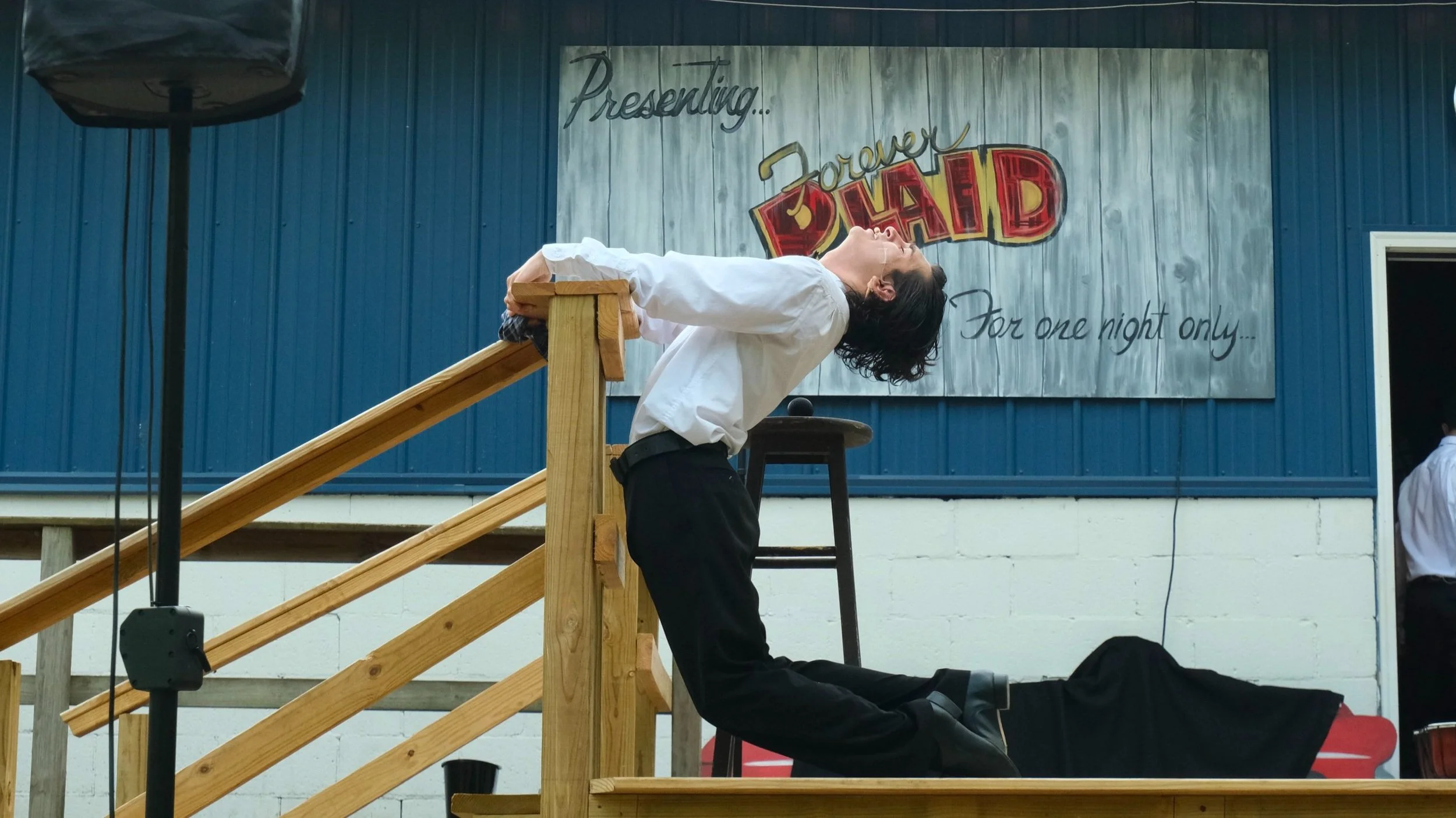 A man in dress pants and a white shirt is kneeling on a stage, arching his back and leaning backward over a wooden railing. Behind him is a sign that reads 'Presenting... Forever Dead For one night only...' in a theater or event setting.