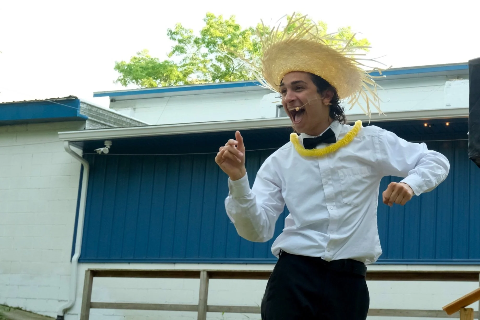 Person wearing a large straw hat, white dress shirt, black bow tie, and a yellow lei, smiling and dancing outdoors in front of a blue building with trees in the background.