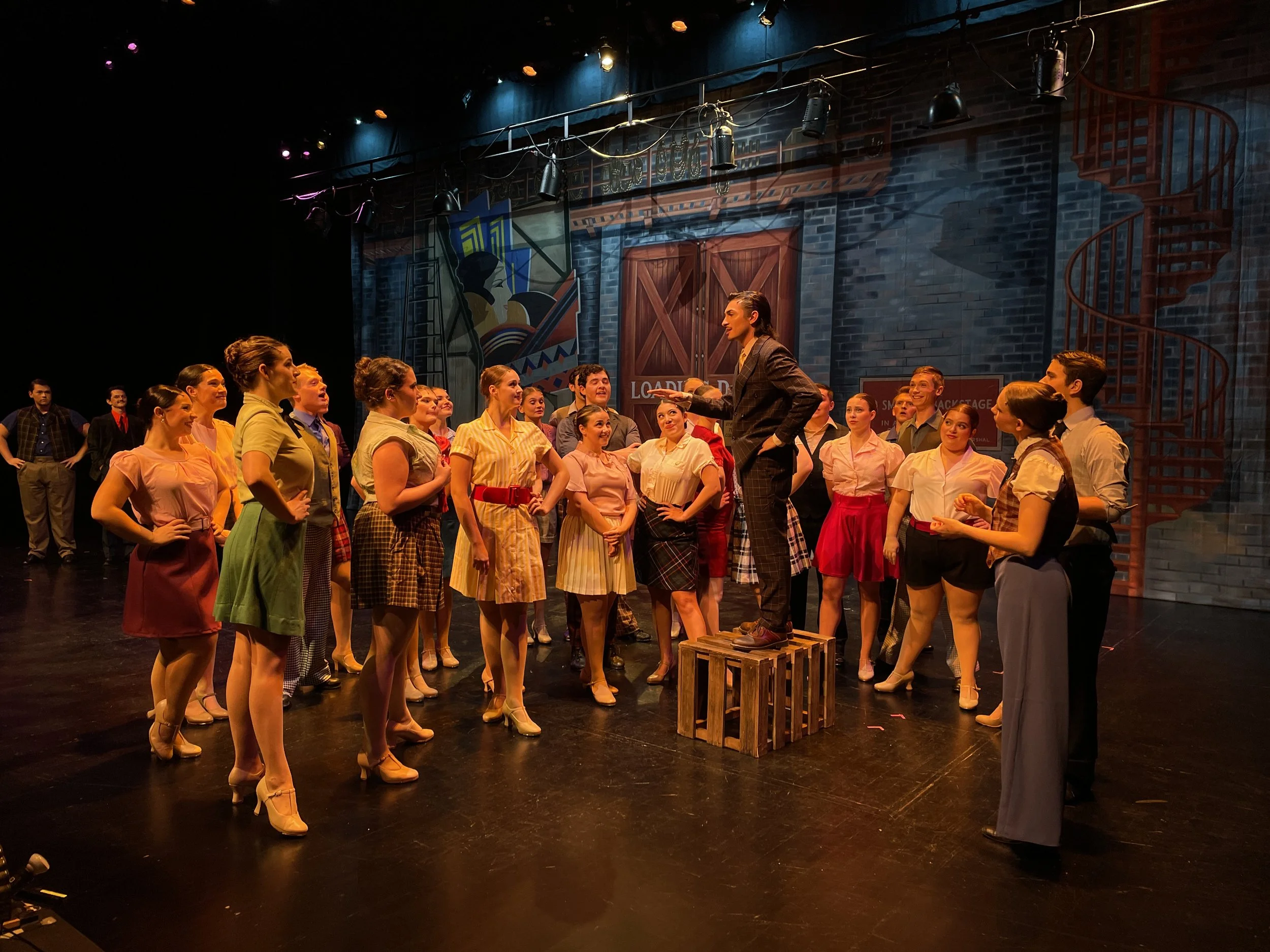 Rehearsal or performance of a stage production depicting a group of actors in vintage attire, with a woman standing on crates presenting to others against a theatrical set background.