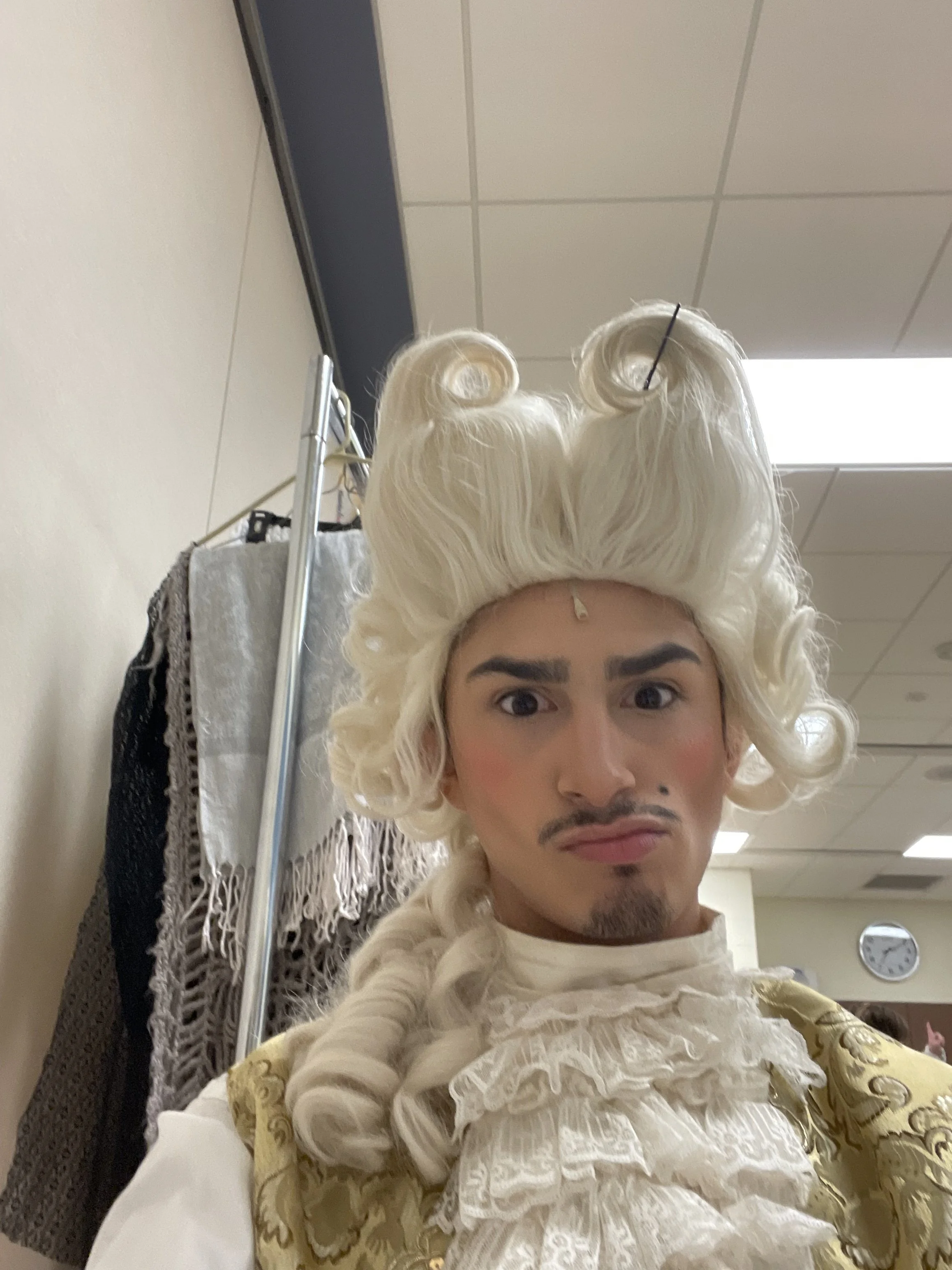 Person dressed as a historical figure with a white powdered wig, elaborate gold and cream outfit, and makeup, taking a selfie in a room with a beige wall, clothing rack, and clock.