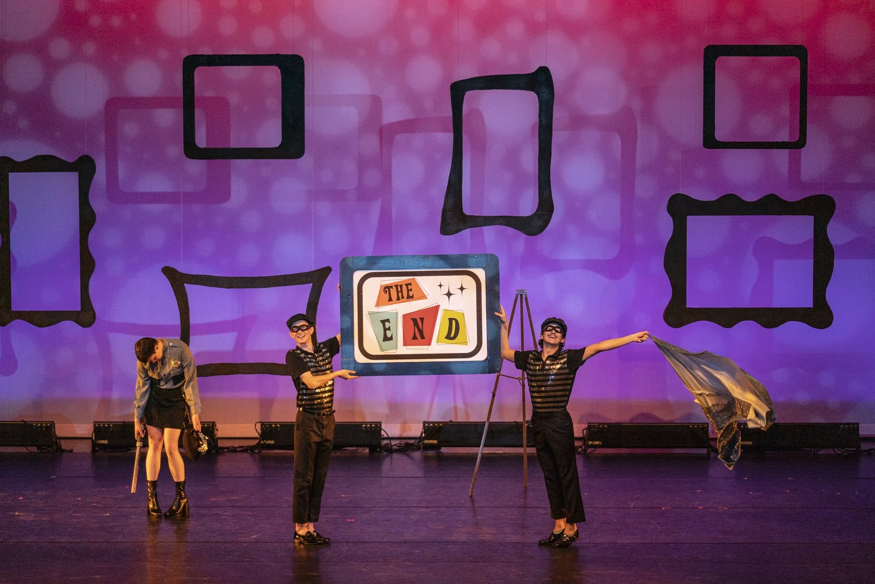 Four performers on stage with a purple backdrop featuring black outline picture frames. Two performers are holding a large sign that says 'THE END,' and the other two are acting out a scene, one holding an umbrella and the other adjusting costume acc