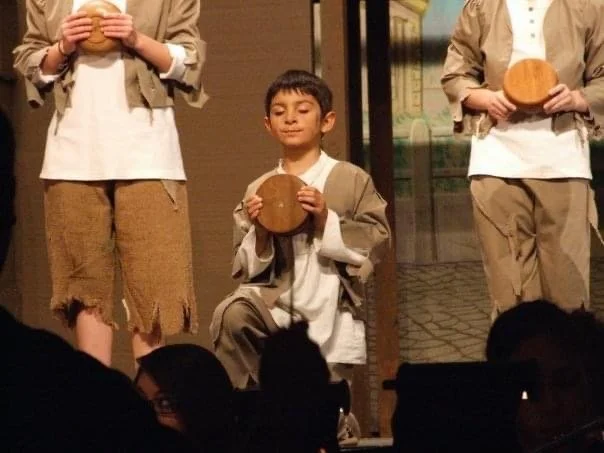 A young boy on stage holding a round wooden object, with two adults partially visible holding similar objects, during a theatrical or school performance.