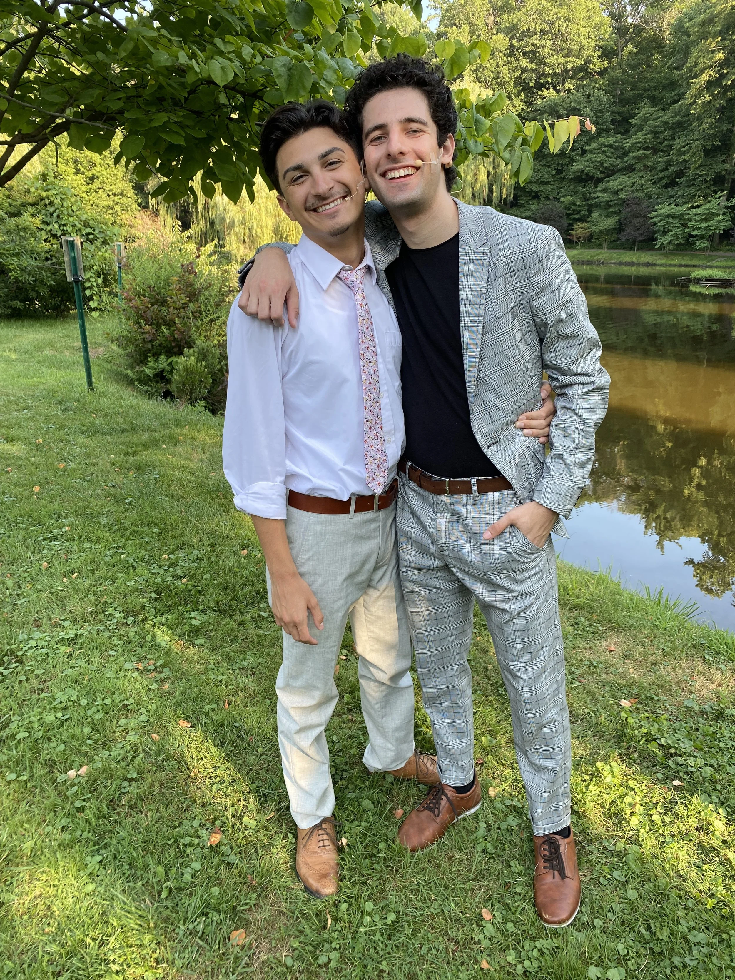 Two young men standing on grass near a pond, smiling and hugging each other, surrounded by green trees.