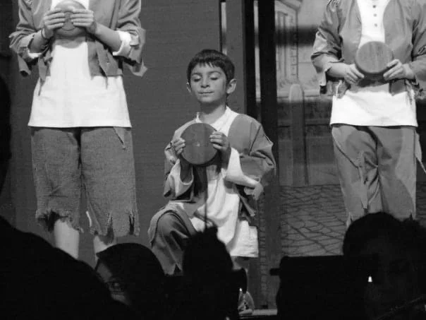 A young boy kneeling on stage holding a round object, with two other people standing around him also holding round objects.