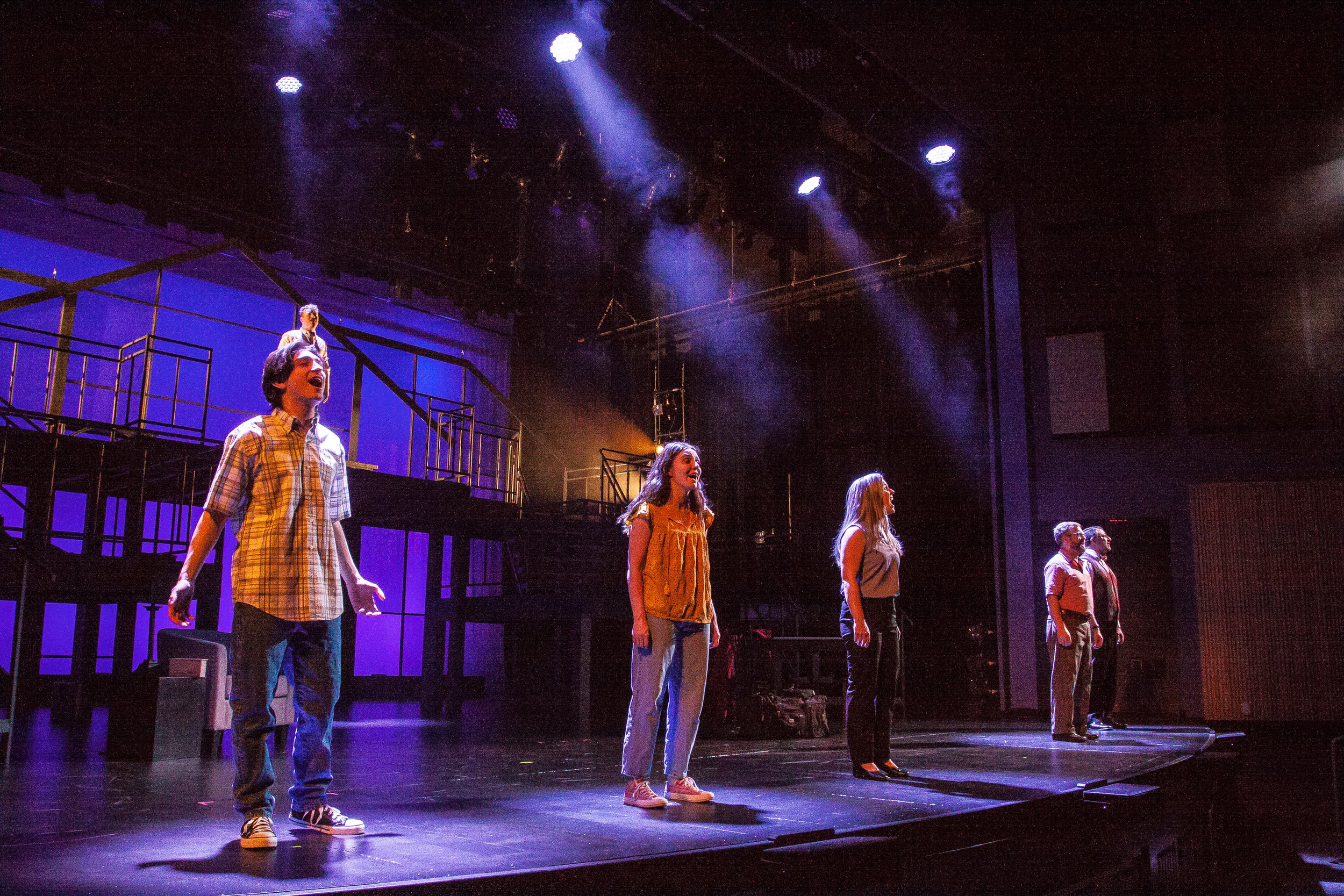 Actors on stage during a theater rehearsal or performance, with three women and two men standing in a line, illuminated by stage lighting, with a set in the background.