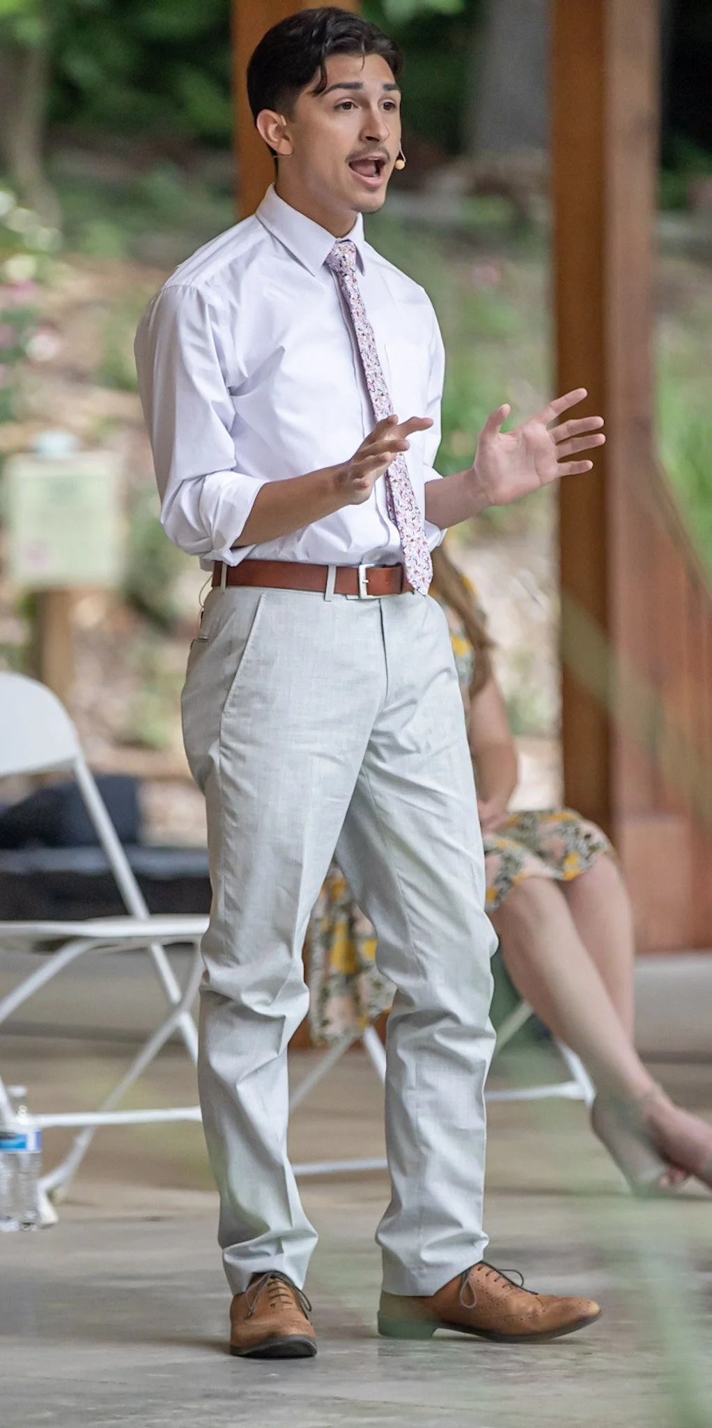 A man with dark hair and a mustache, wearing a white shirt, patterned tie, beige pants, and brown shoes, speaking or giving a presentation outdoors with a blurred woman sitting behind him.