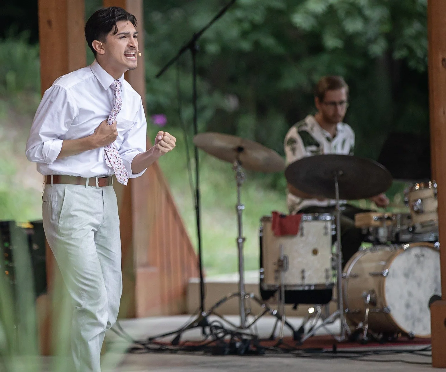 A man in a white shirt and light-colored pants passionately singing into a microphone, with a drummer playing drums on a stage outdoors in the background.