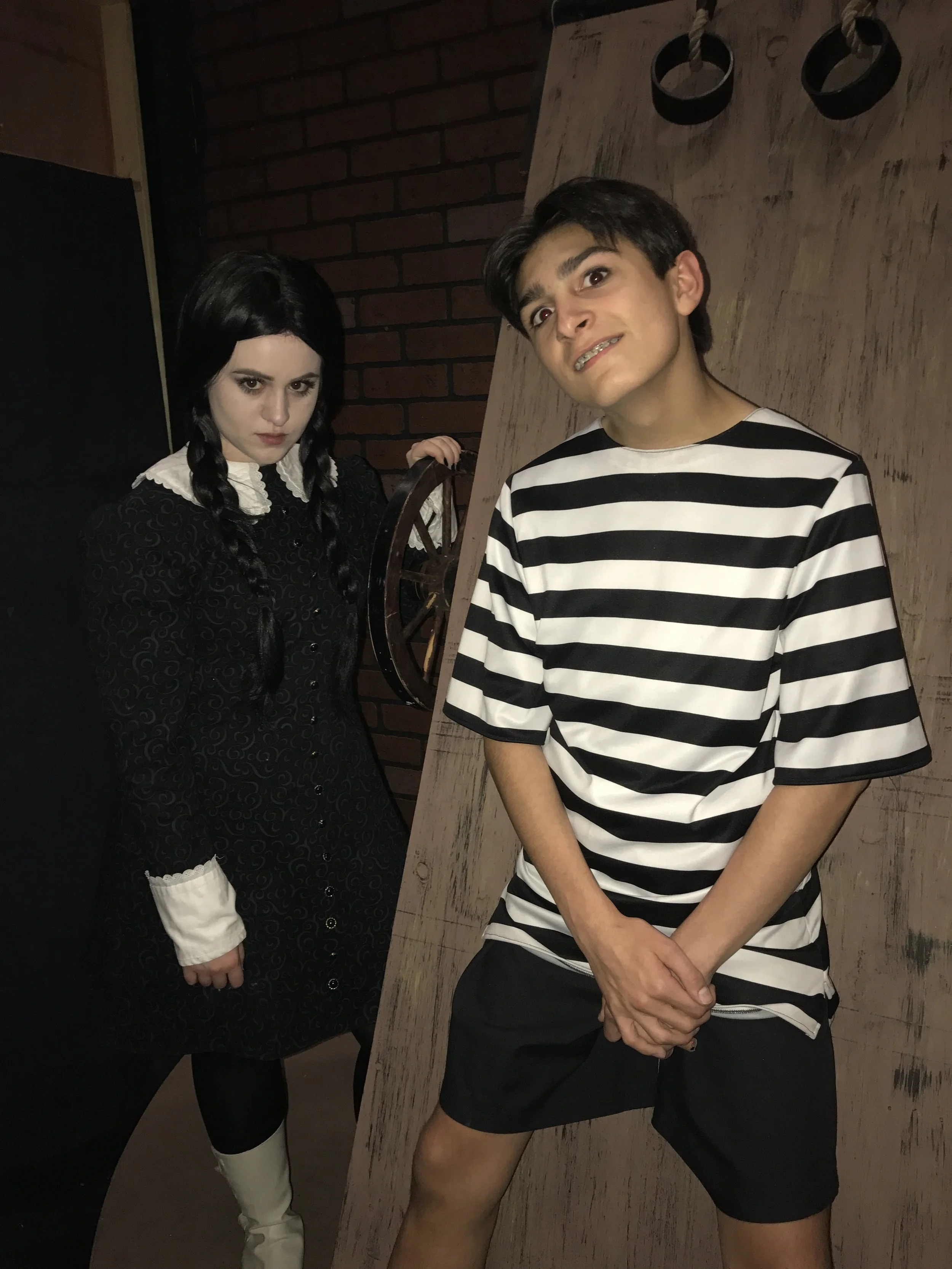 A young man in a black and white striped shirt and shorts posing with a playful expression, standing in front of a wooden wall. A girl with long dark hair in braids, dressed in a black dress with white cuffs and collar, stands behind him, holding a w