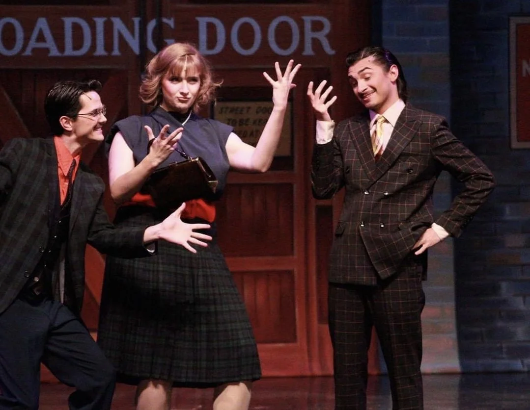 Three actors on stage performing a scene, one woman in the middle with crossed arms and an expressive face, a woman on the right gesturing with her hand, and a woman on the left smiling and leaning in.
