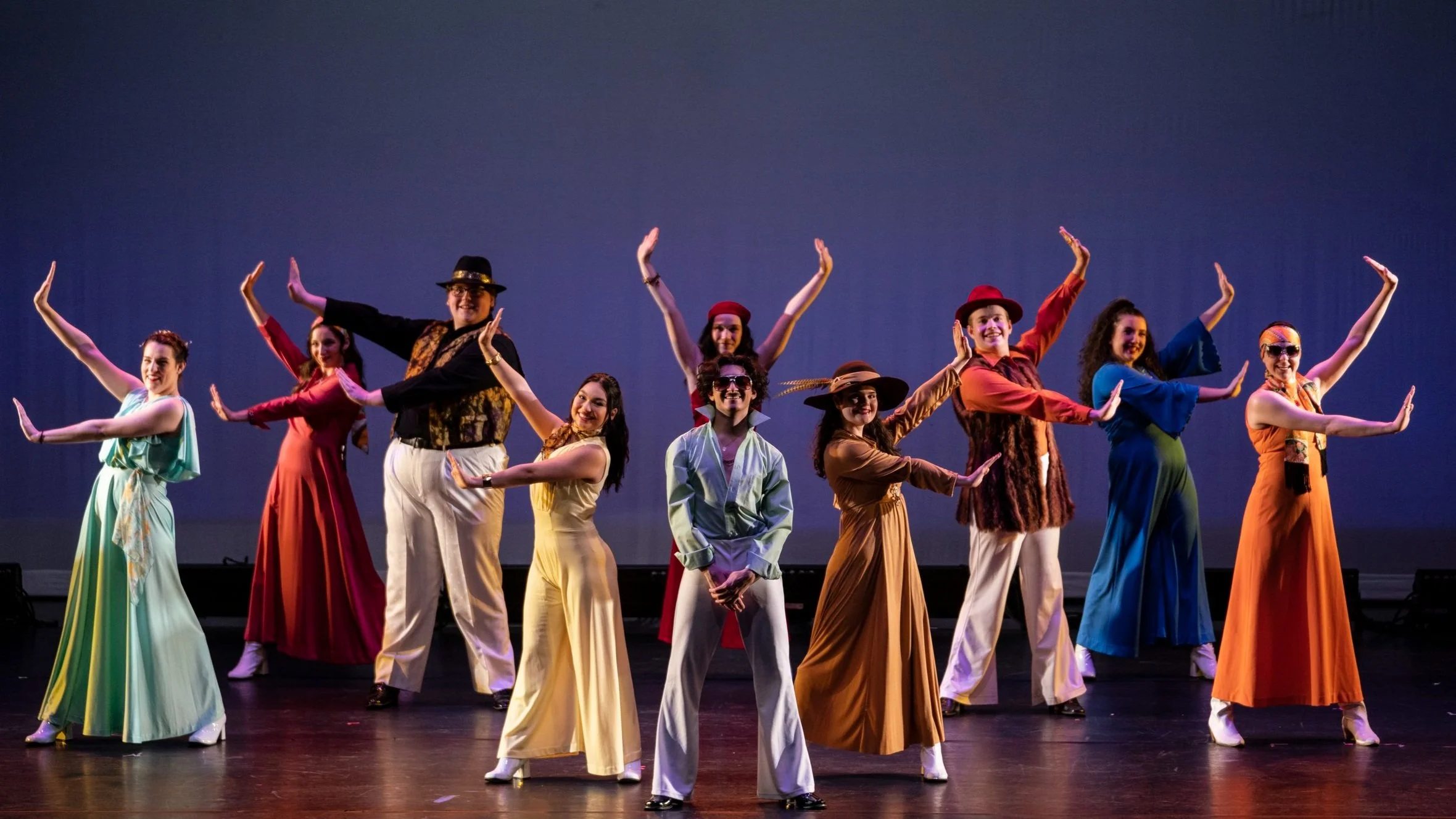 The image shows a diverse group of eleven performers on stage, dressed in colorful vintage costumes, striking a synchronized pose during a dance routine with a plain blue backdrop.