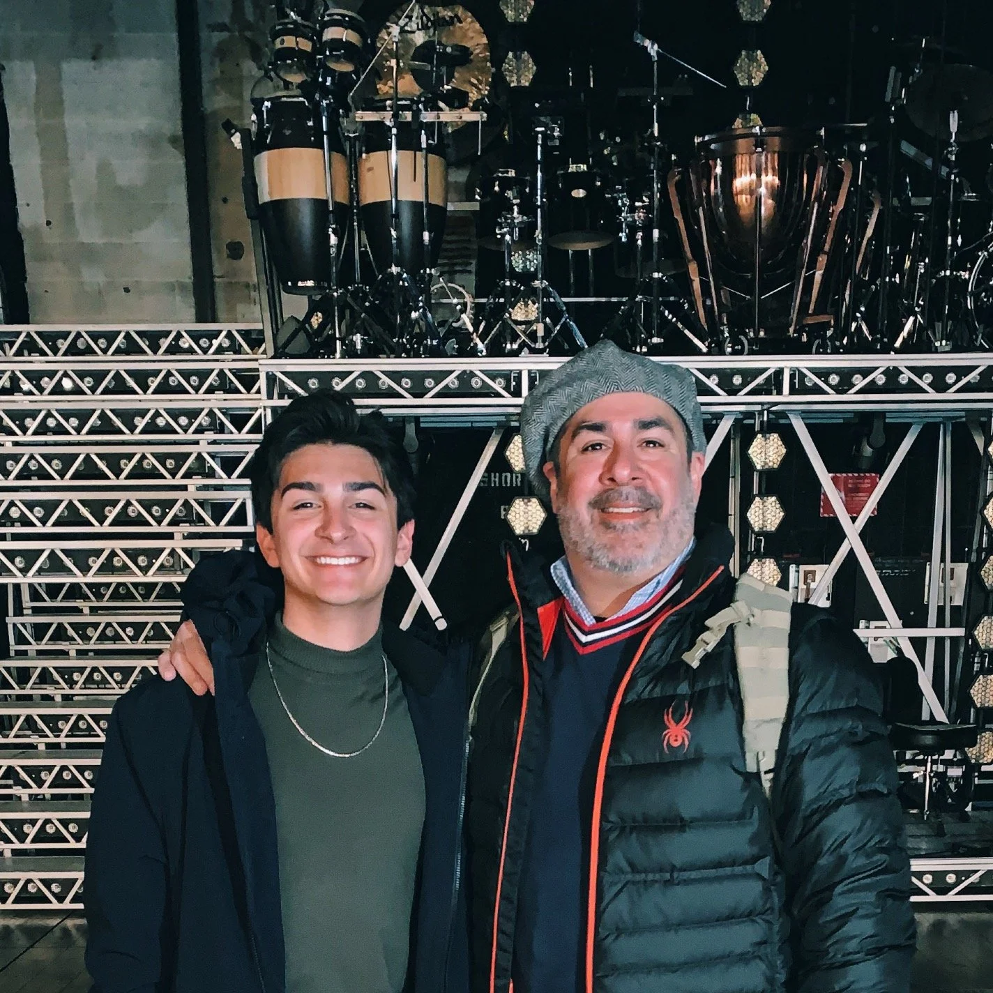 Two men smiling, with one having his arm around the other, standing in front of a musical stage with drums.
